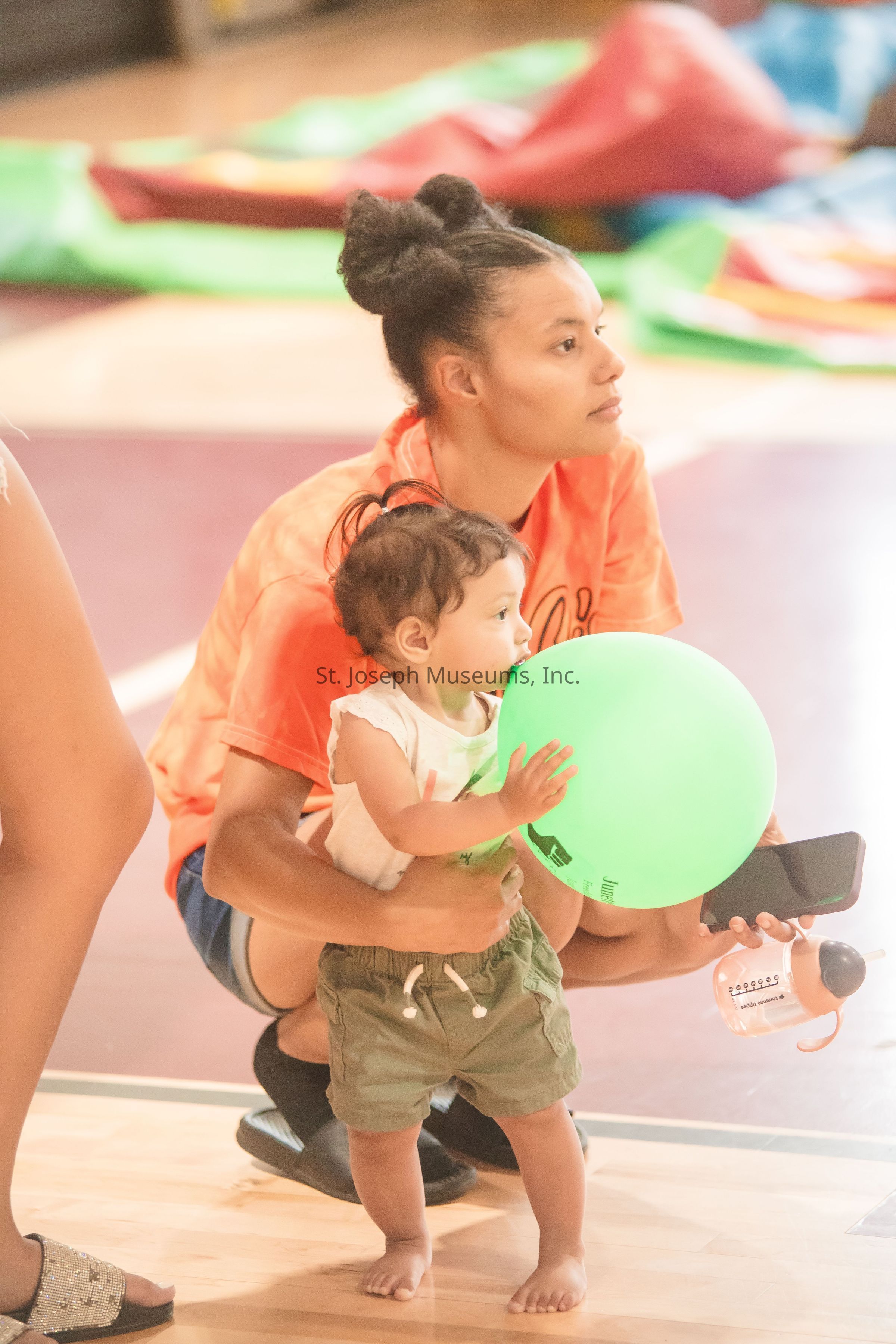 In a colorful indoor setting, a young child with curly hair stands on a wooden floor, intently holding a large green balloon, while being gently supported by an adult who is crouching beside them. The adult, wearing an orange top, looks onward, holding a smartphone in one hand. A small, orange-lidded bottle is visible next to them on the floor. The background is softly blurred with vibrant, multicolored inflatable play structures creating a lively atmosphere. The image captures a moment of attentive care and focused playfulness, depicting a warm interaction between the child and the adult, surrounded by a bright and playful environment.