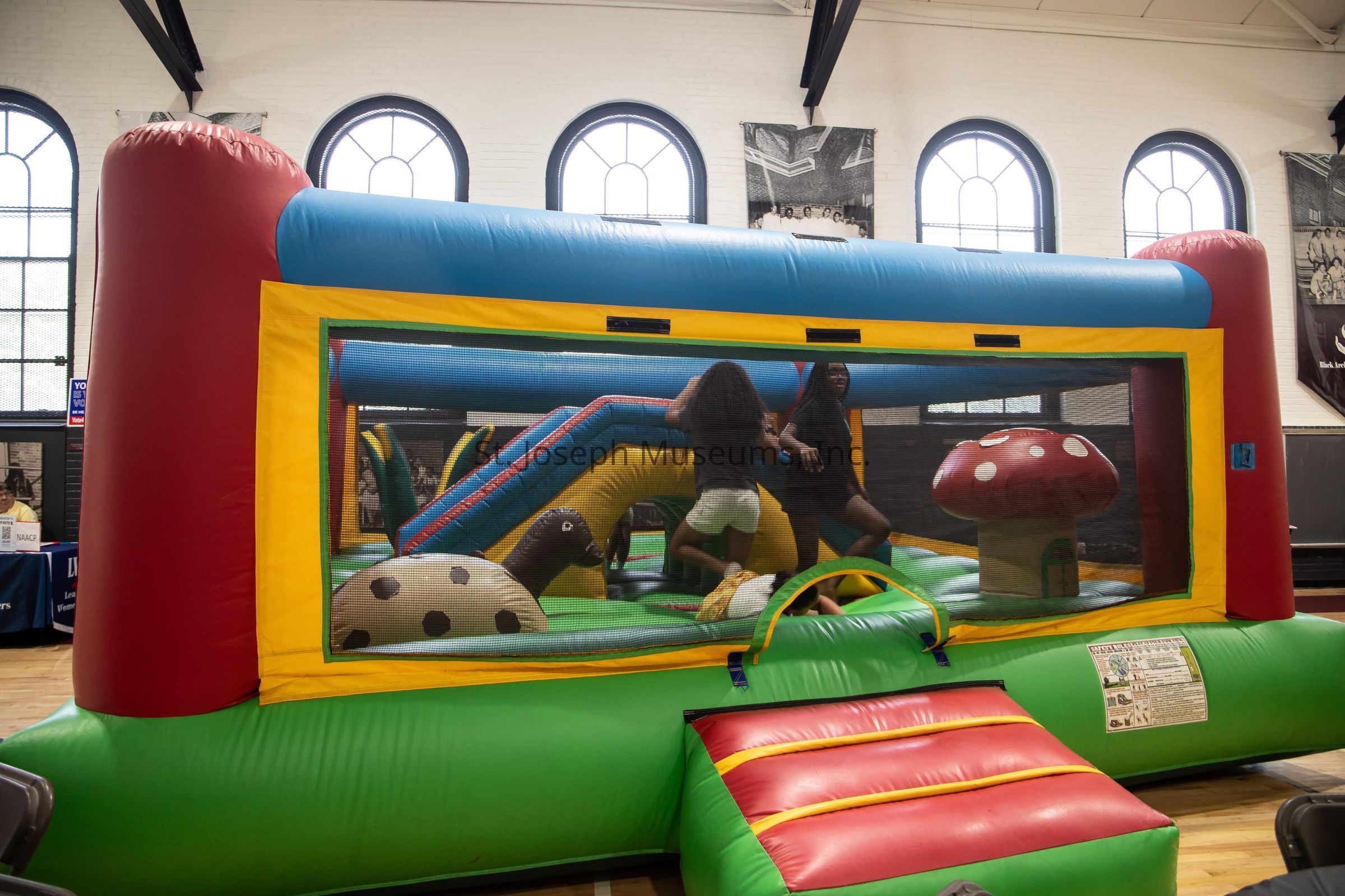 The image shows an indoor play area featuring a colorful inflatable bounce house designed for children. The bounce house is vibrant, with red, blue, yellow, and green sections, and has a transparent mesh on one side to allow visibility inside. Two children are playing inside, surrounded by inflatable structures resembling playful designs like a spotted mushroom and a slide. The bounce house is set up in a large room with high, arched windows that let in natural light. The floor is wooden, suggesting it's in a gymnasium or similar indoor facility. In the background, a table with people seated and informational displays can be observed, indicating perhaps a fair or community event in progress.