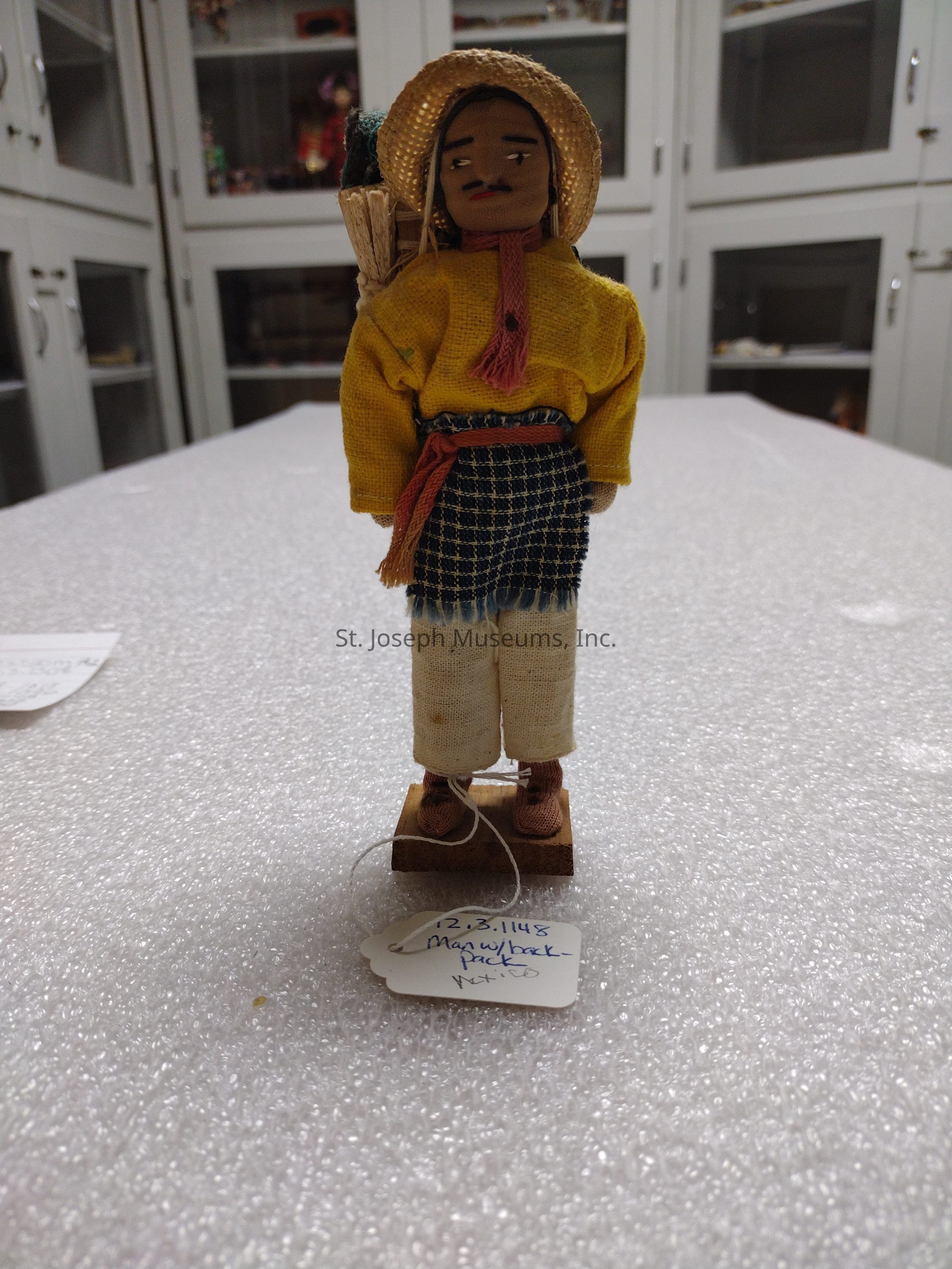 The image shows a handcrafted figurine representing a man dressed in traditional attire standing upright on a textured surface. He is wearing a bright yellow shirt and a woven skirt with a checked pattern, along with light-colored pants and brown shoes. On his head, he has a straw hat partially covering his head, and his expression is accentuated with facial features such as painted eyes and a mustache. Attached to the figurine is a string with a tag that reads, "12.3.1148 Man with backpack" in blue ink. The man carries a detailed woven basket backpack, giving a rustic, handmade charm to the overall appearance. The setting appears to be inside a room with glass-front cabinets, which likely serve as display cases for various artifacts or collectibles, providing a backdrop of cultural and historical interest.