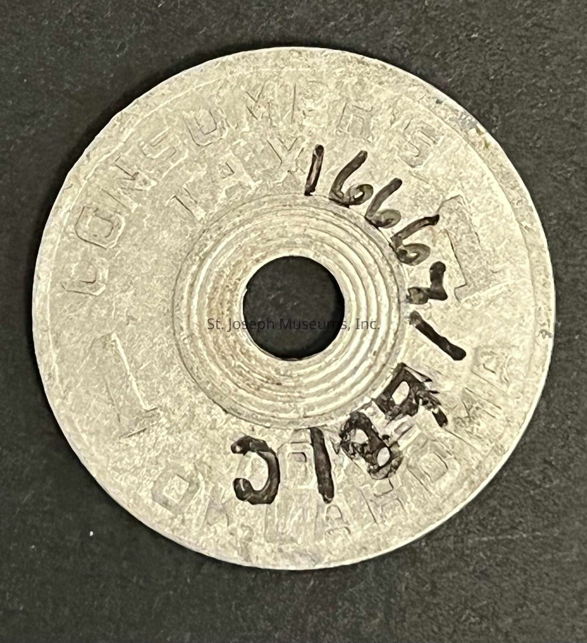 The image shows a round, metal token with a central hole. The surface of the token has engraved text that reads "CONSUMER'S TAX" and "PRIVATE." The token is placed against a dark background, highlighting its metallic texture and any wear it has sustained over time. The markings, both engraved and hand-written, suggest it was used for a specific purpose, possibly as a form of voucher or tax-related identifier.
