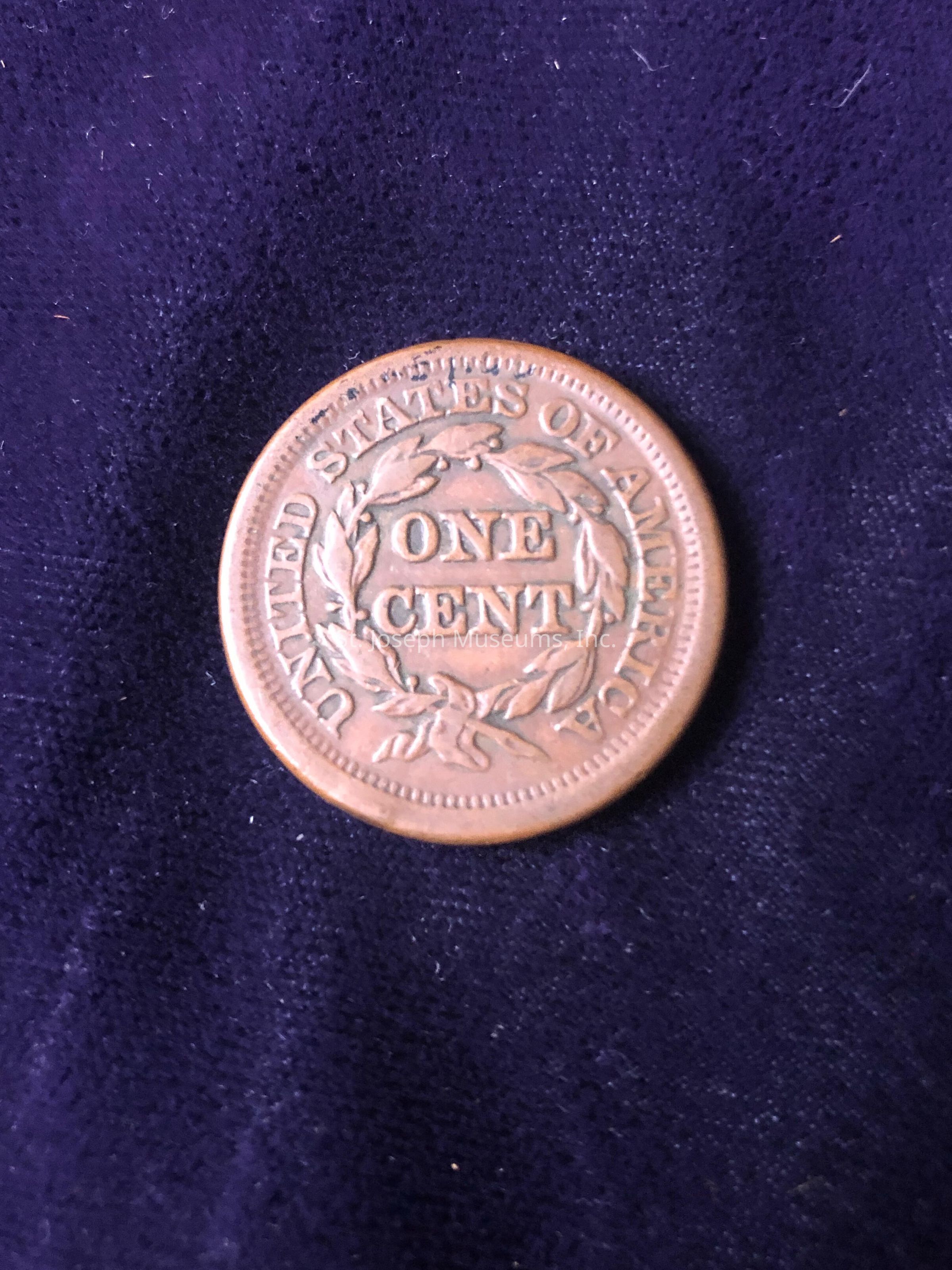 The coin's design features the words “UNITED STATES OF AMERICA” along the edge and "ONE CENT" prominently in the center, encircled by a decorative wreath. The coin appears to be worn, indicating its age and potential historical value. The dark fabric beneath the coin provides contrast, making the details on the coin surface more visible. The overall composition draws attention to the coin, suggesting it could be of numismatic interest to collectors or enthusiasts.