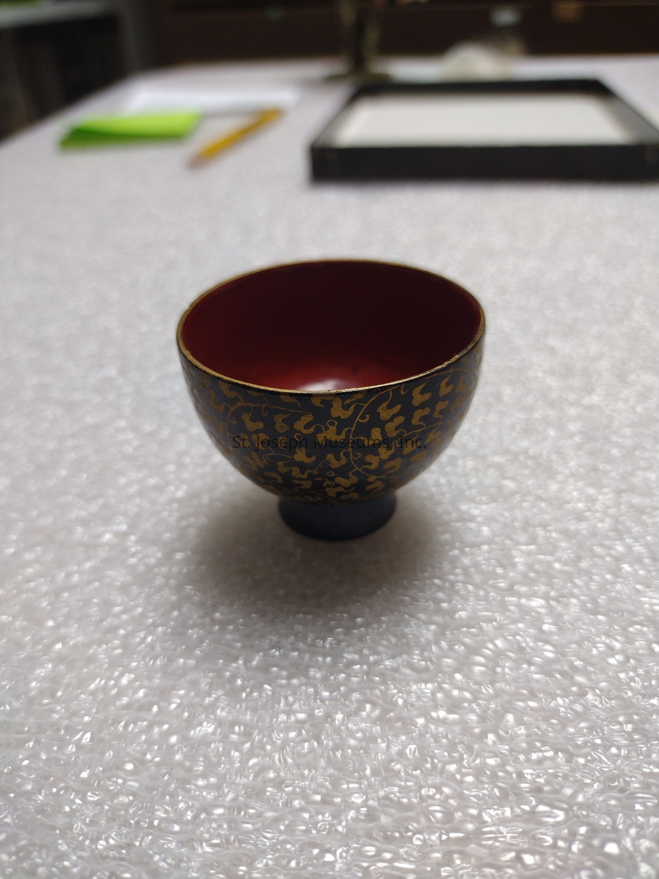 The image depicts a small, intricately designed bowl displayed on a textured surface, possibly a piece of bubble wrap. The bowl has a dark exterior adorned with elegant gold patterns, reminiscent of traditional or artisanal craftsmanship, while its interior is a deep red color. Positioned in a well-lit environment, the bowl is the central focus, surrounded by a blurred background that includes an unidentifiable rectangular object, a pencil, and some green paper, all of which suggest a setting involved in art, study, or collection. The subtle details highlight the bowl's elegance and decorative appeal, emphasizing its potential as a handcrafted piece with cultural or historical significance.