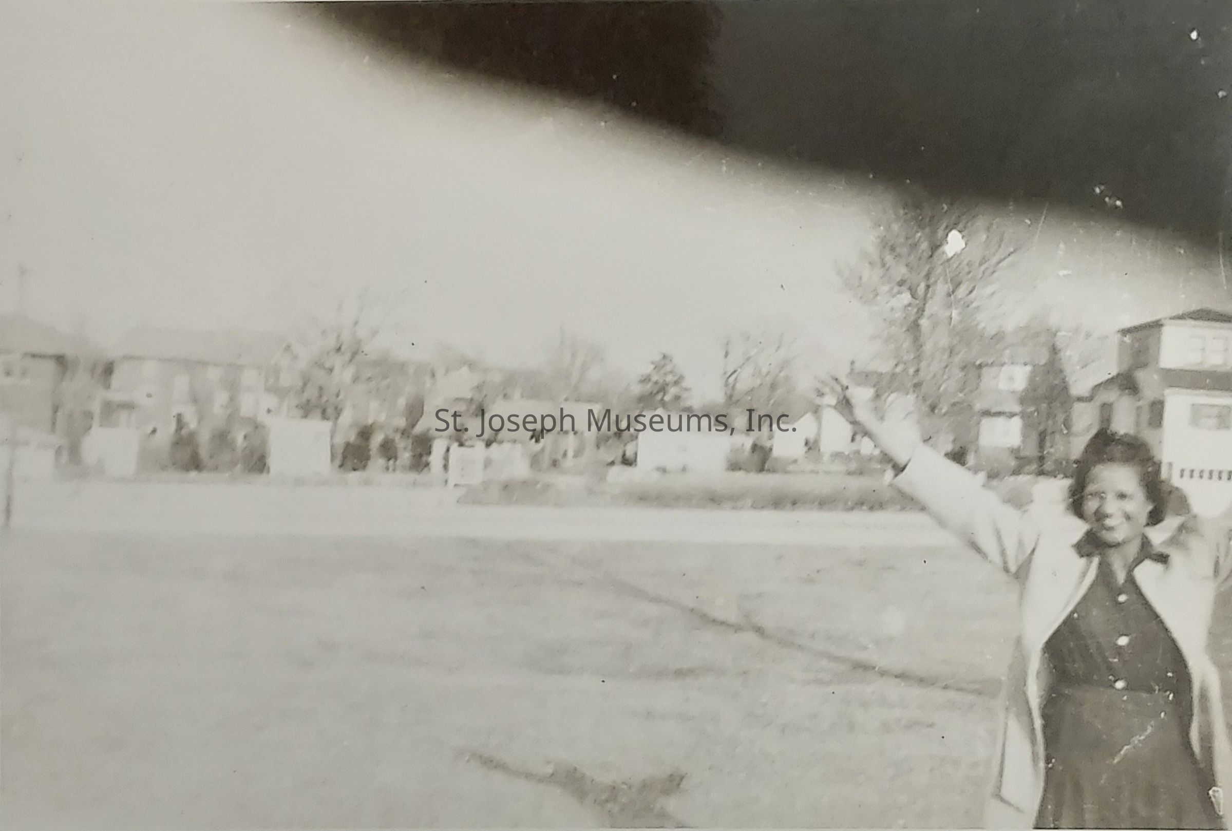 In this black-and-white photograph, a person is standing outdoors, appearing joyful with arms raised high in a celebratory manner, while wearing a coat over a dress. The background reveals a glimpse of suburban architecture, featuring a row of houses with trees that are likely leafless, suggesting either late autumn or early spring. The ground is a grassy expanse with patches that may be slightly uneven, while overhead, the photo appears to have an overexposed or light-leaked portion, adding a vintage or aged effect to the image. The person's expression and posture convey a sense of happiness or triumph, capturing a candid moment within a quiet, residential neighborhood.