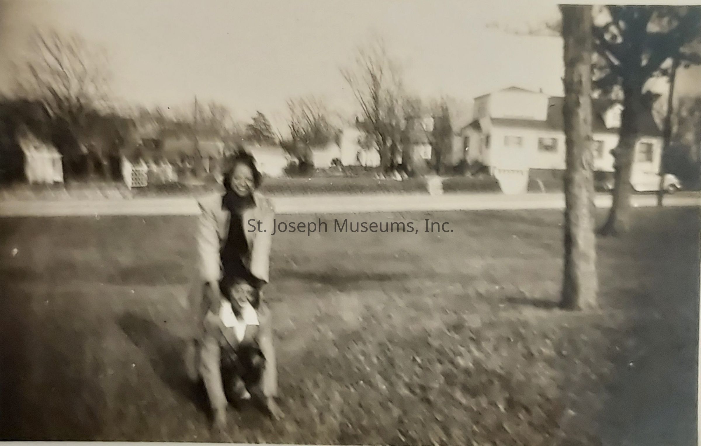 The black and white image captures a heartwarming moment between two people in an outdoor setting; a woman, standing behind, places her hands gently on the shoulders of a child who crouches playfully in front of her, both radiating joy as they engage in a delightful game; the background reveals a peaceful suburban neighborhood lined with classic mid-century houses, bare trees, and an open grassy area suggesting the season could be late autumn or early spring, while the slightly blurred and nostalgic appearance of the photograph evokes a sense of timelessness and familial warmth, characteristic of cherished memories from the past.