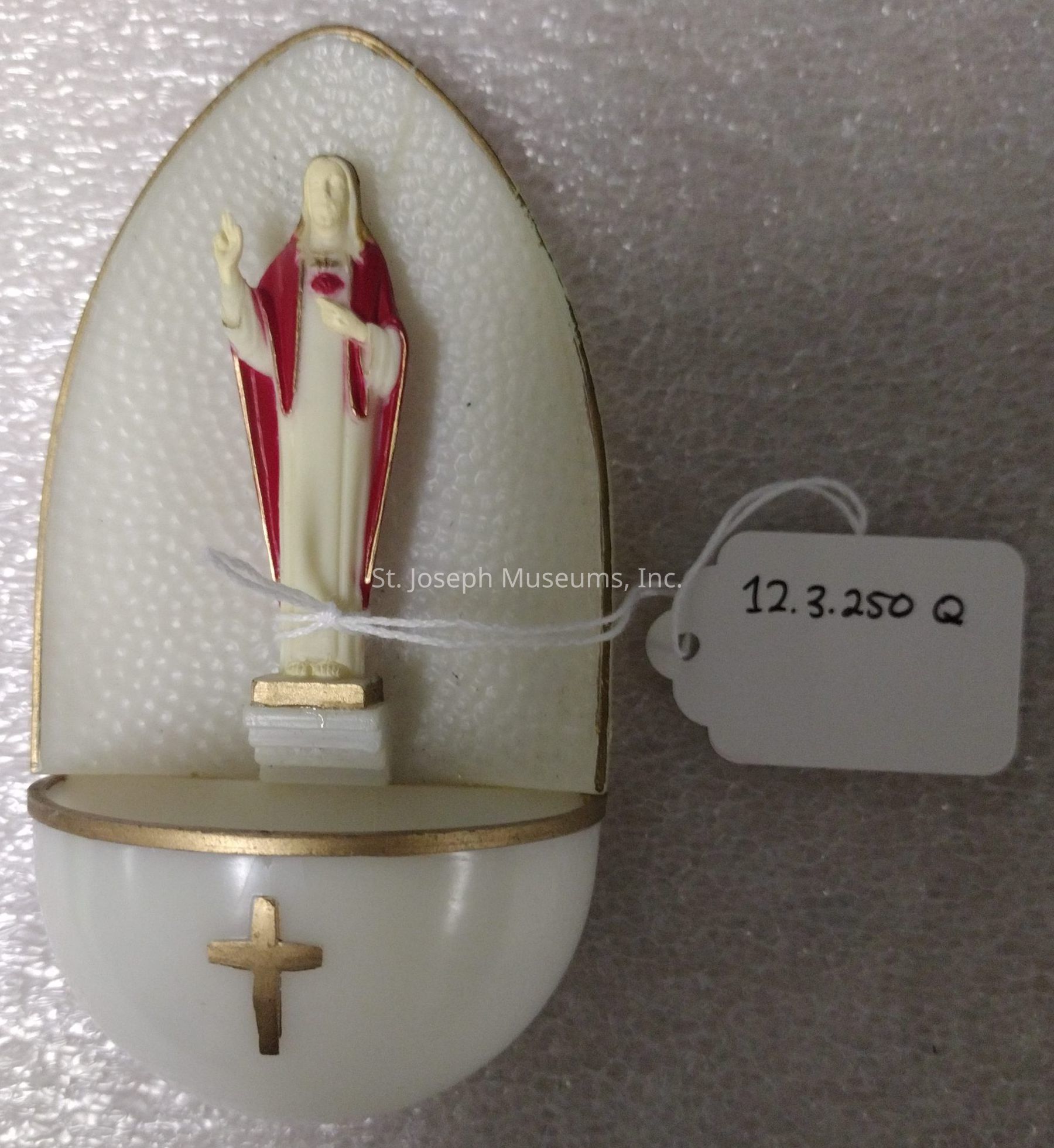The image depicts a religious wall-mounted holy water font, featuring a small statue of a robed figure with long hair in a prayer or blessing gesture, set against a textured, oval-shaped background with gold trim. The bottom section is a semi-circular container for holding holy water, embellished with a gold cross on its front. Attached to the font is a small white tag with the inscription "12.3.250 Q," suggesting that it may be part of a collection or inventory. The entire piece rests on a speckled, textured surface, likely for display or storage purposes.