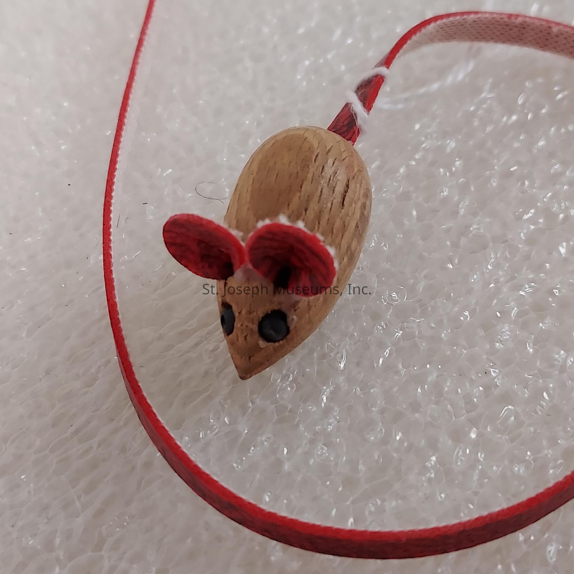 The image features a small, wooden, mouse-shaped ornament with two large pink ears and two black eyes, giving it a cute and whimsical appearance. The mouse has a long, thin red ribbon attached at the back, resembling a tail, which is artistically looped above a textured, white, bubbly surface, possibly a type of foam or packaging material, providing a contrasting background that highlights the natural wood grain and bright colors of the mouse figurine's features. The simplicity and design suggest it could be a handcrafted decoration or toy, evoking a sense of charm and playfulness.
