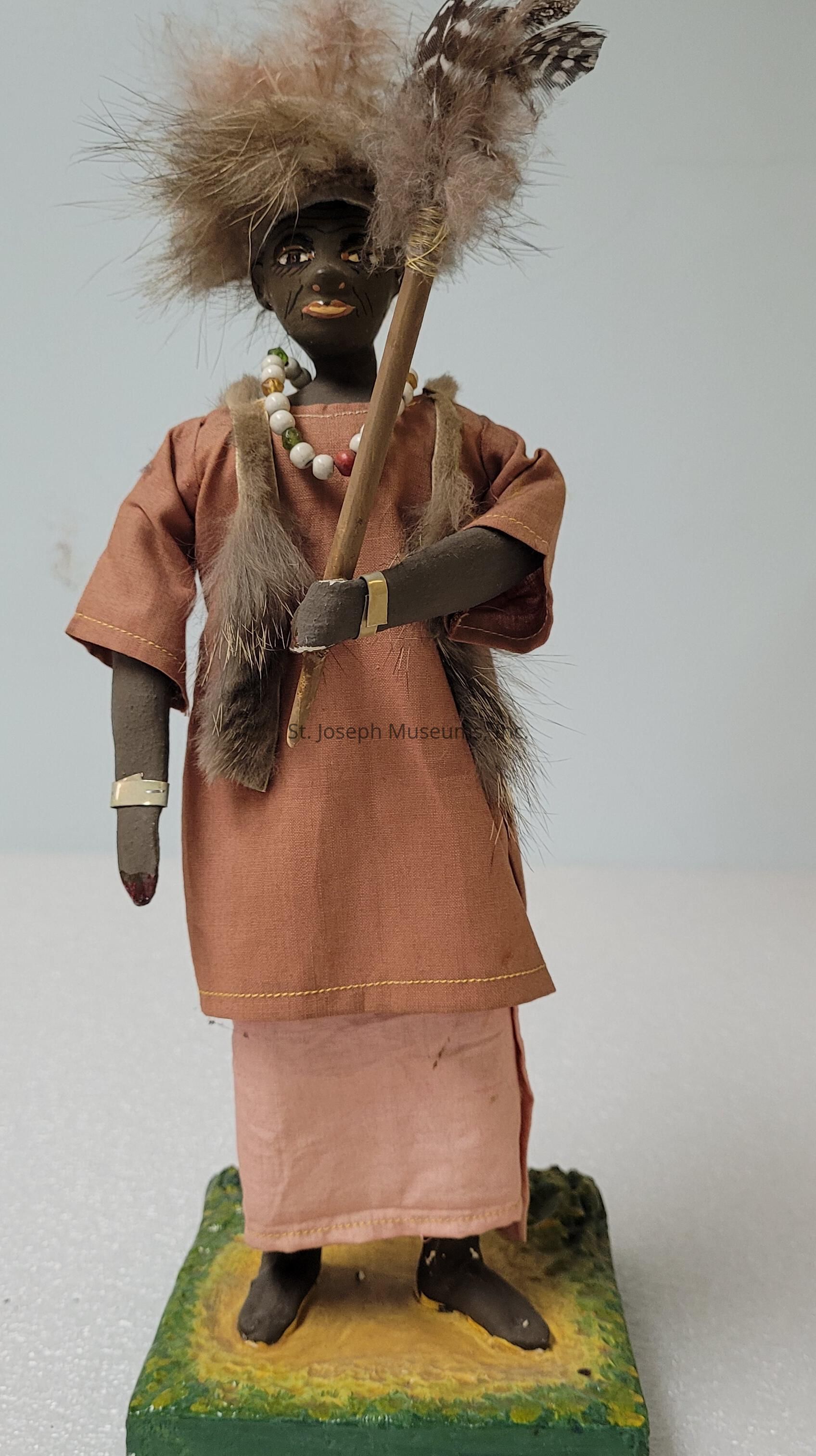 The image features a handcrafted figurine depicting a person dressed in a traditional outfit consisting of a brown top and a pink lower garment. The figure's attire is adorned with fur-like details on the shoulders and accessorized with a necklace of colorful beads. Its hair is styled with feather-like material that complements the feathery staff it holds, all of which give it an earthy, tribal appearance. The hands and feet of the figure display dark paint, adding to the distinct contrast of its overall aesthetic. The figurine stands on a square base with simulated grass and earth textures, enhancing the natural theme. This artistic representation showcases a creative blend of materials and textures, including fabric, fur, beads, and paint, all cohesively assembled to convey a cultural and artistic expression.