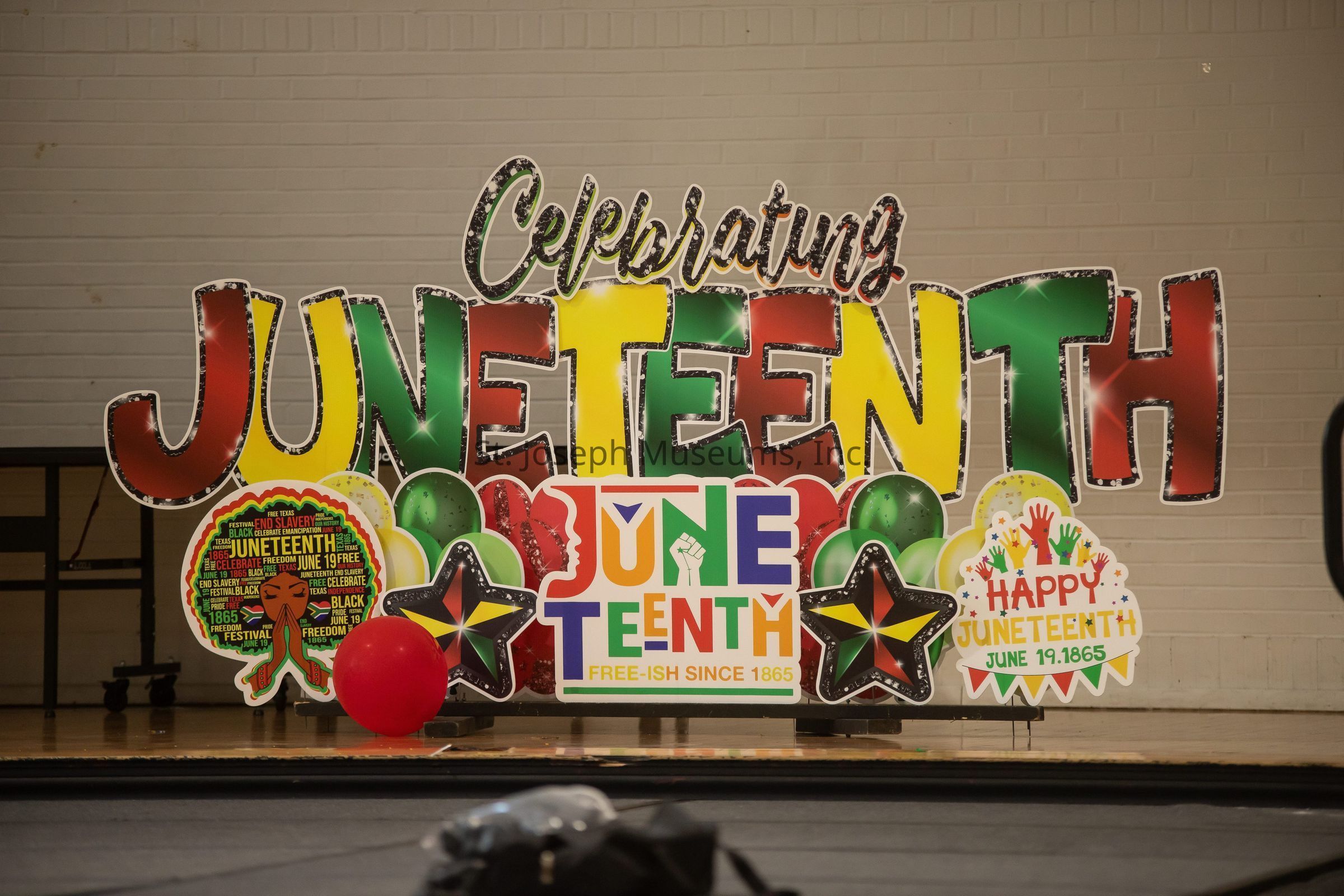 The image displays an elaborate setup celebrating Juneteenth, featuring large, colorful cutouts and decorations. The word "Juneteenth" is prominently highlighted, surrounded by vibrant red, green, and yellow colors. The phrase "Celebrating Juneteenth" is written above in a festive font, and additional elements include stars, balloons, and thematic graphics, such as clenched fists and profile silhouettes, symbolizing freedom, unity, and pride. The messages "Free-ish Since 1865" and "Happy Juneteenth, June 19, 1865" emphasize the historical significance of the event. A red balloon is placed in front, adding to the celebratory atmosphere.