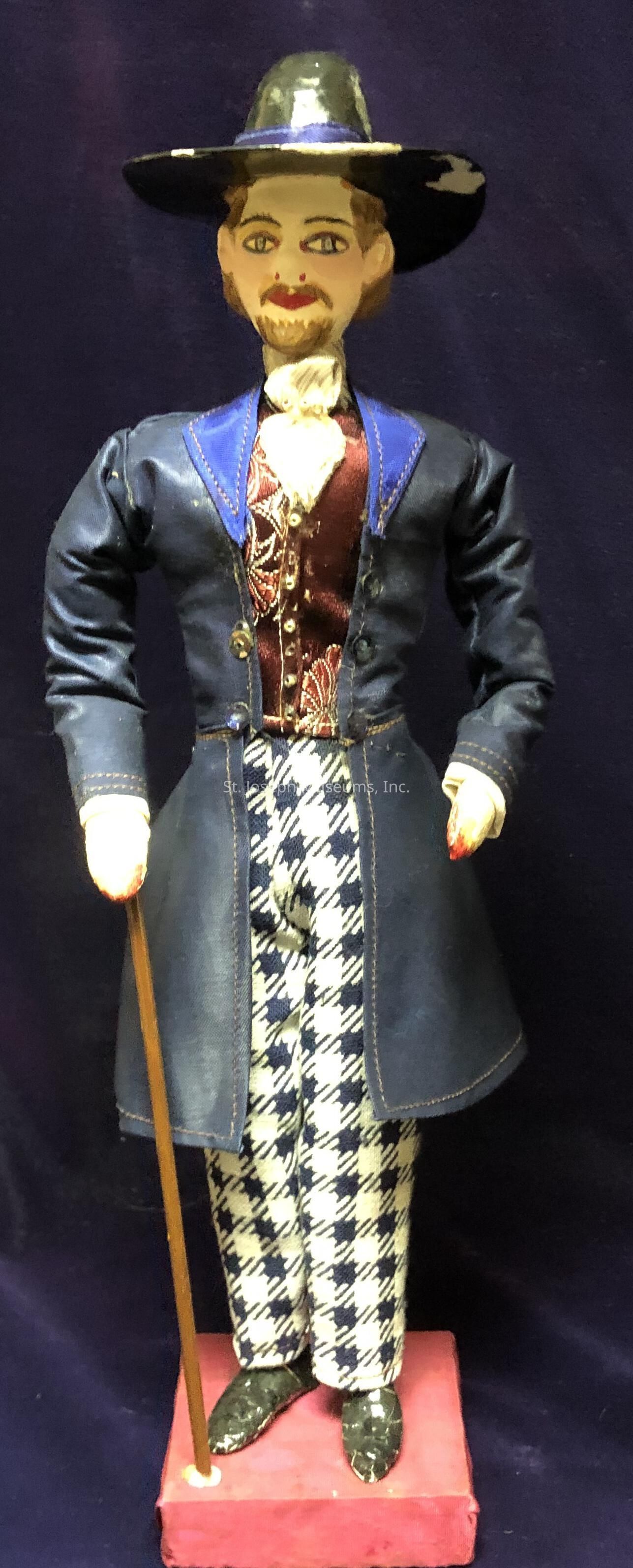 The image shows a vintage puppet or marionette dressed in a distinctive, formal outfit, standing on a red platform against a dark background. The figure wears a blue suit with detailed stitching, adorned with a maroon vest featuring ornate patterns and a white cravat at the neck. Its pants display a black and white checkered pattern, adding contrast to the ensemble. A black hat with a wide brim sits atop the puppet's head, complementing its stylized painted face, complete with visible eyes and a neatly groomed beard and mustache. The puppet holds a wooden cane in its left hand, enhancing its dignified appearance.