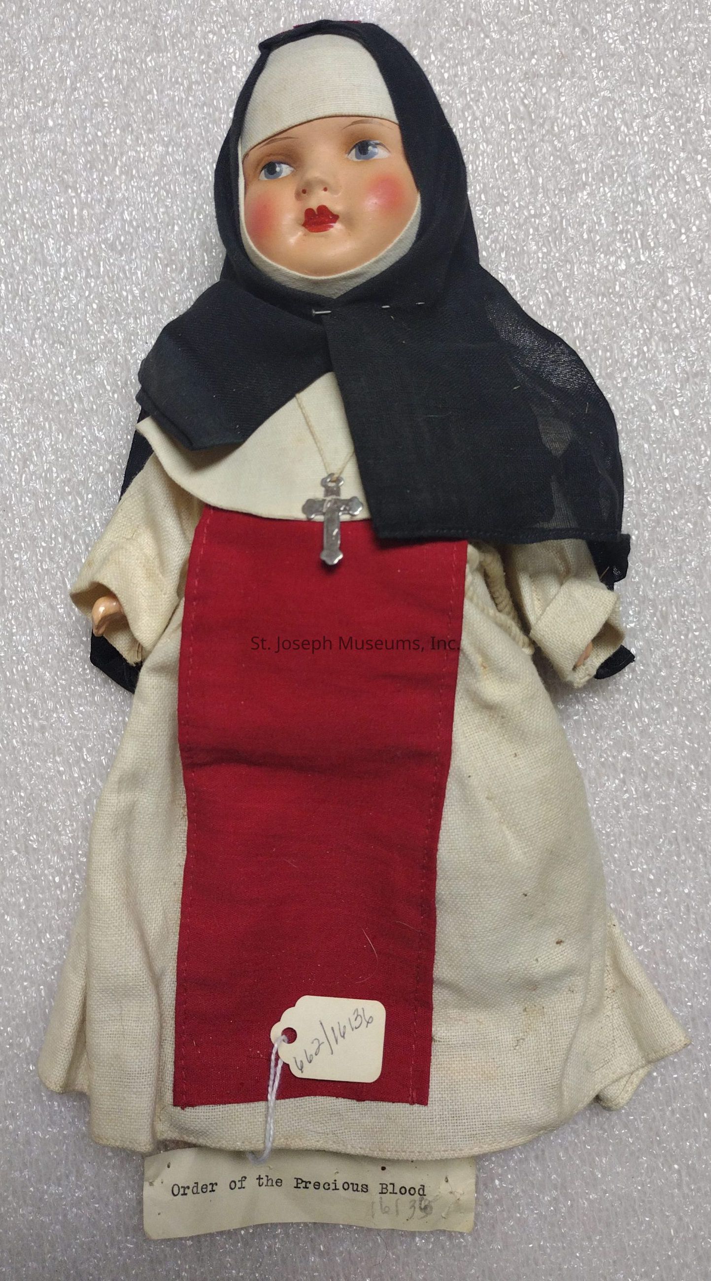 The image shows a vintage doll dressed in a nun's habit, including a black veil and a white headdress. The doll's face is painted with rosy cheeks and red lips, and it wears a cross on a chain around its neck. The habit includes a prominent red panel down the front of the white robe. Attached to the doll is a tag with numbers and a typed note stating "Order of the Precious Blood." The background is a textured, light-colored surface that resembles a fabric or cushioning material, which highlights the intricate details of the doll's attire and adds context to its historical and religious significance.