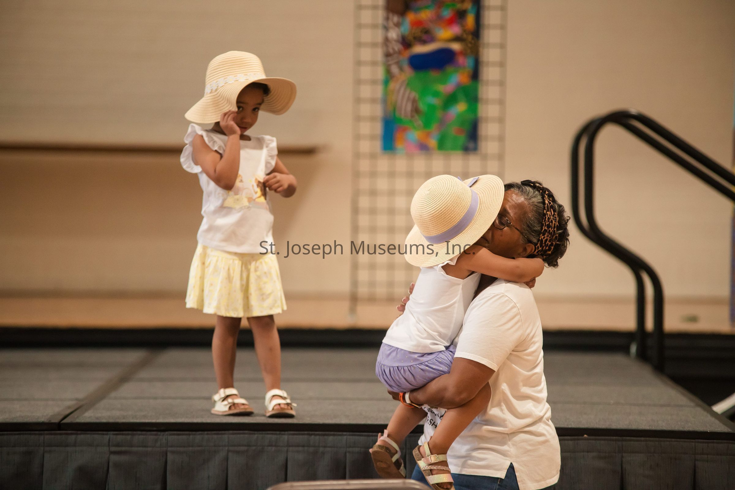The image depicts a touching scene where a woman affectionately embraces and lifts a young child who is wearing a straw hat, in her arms, while another child, also wearing a straw hat and dressed in a yellow skirt and white top, stands nearby on a stage, observing the interaction with a thoughtful expression. The background features a colorful abstract painting and a staircase, suggesting the setting is a community or cultural event. The heartfelt moment is highlighted by the tender gesture of the woman and her joyful interaction with the children, creating a warm and loving atmosphere.