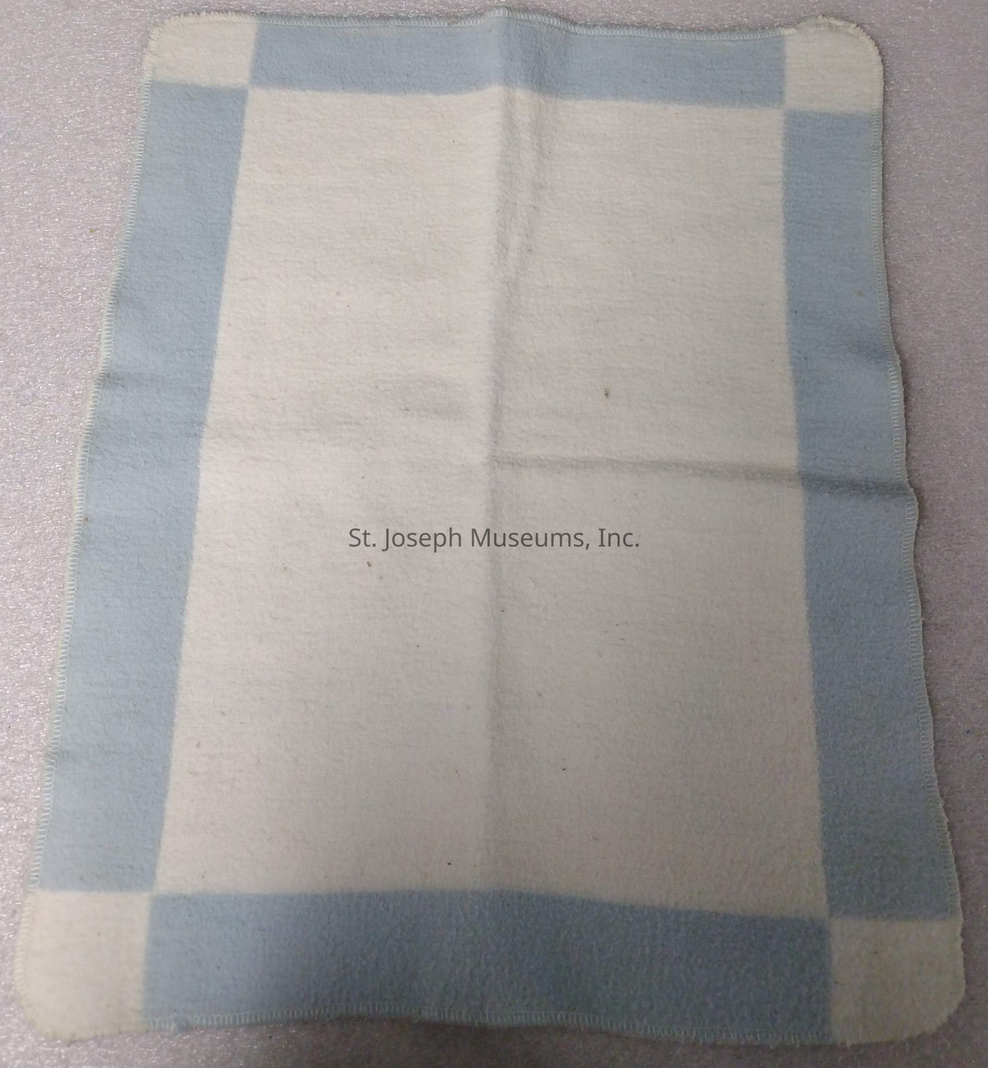 The image shows a rectangular woolen blanket laid out on a textured surface. The blanket features a simple yet elegant design, primarily consisting of a light cream color with a contrasting, soft blue border. This blue border frames the blanket, creating a visual boundary that enhances its aesthetic appeal. The fabric appears to be of a cozy and warm material, suggesting comfort and functionality. Subtle variations in texture are visible across the blanket's surface, indicative of its material's softness and plush quality. The edges of the blanket are finely stitched, ensuring durability and a neat finish. The overall appearance of the blanket is classic and timeless, making it suitable for various decor styles and functional uses, such as providing warmth or adding a layer of comfort to a setting. Its gentle color palette suggests it might complement a wide range of interior designs, adding a touch of understated elegance and comfort to the environment in which it is used.