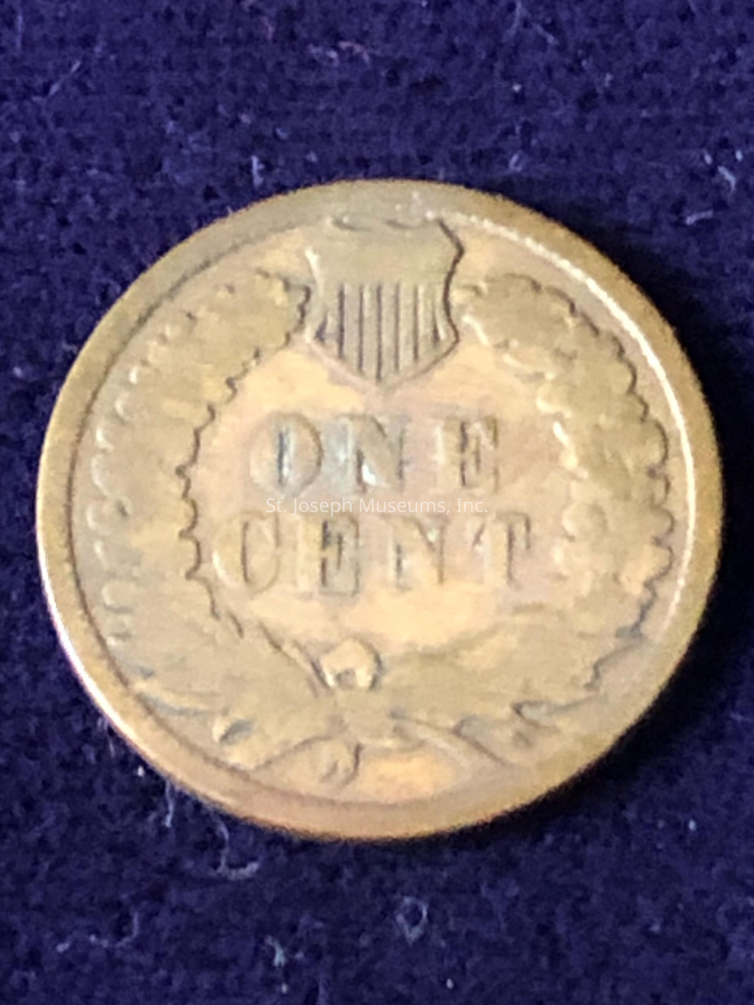 The coin is round, with a shield emblem at the top and the words "ONE CENT" prominently displayed in the center. Surrounding the text is a wreath design that adds detail to the coin's surface. The overall color of the coin suggests that it is made of a metal with a bronze or copper-like hue, which is characteristic of older coins. The intricate details and the emblem suggest it might be of historical significance, possibly a vintage or antique piece, given its classic design elements.
