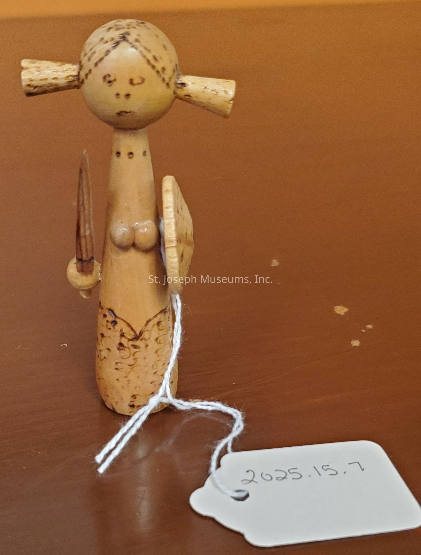 The image depicts a small wooden figurine crafted with intricate details, featuring minimalistic facial features and two buns on its head. The figure is holding a spear and a round shield, suggesting it might represent a warrior or a guard. The body is cylindrical, ornamented with subtle carved patterns. Attached to the figure is a white string with a label, the text on which reads "2025.15.7". It is placed on a smooth, brown surface, creating a warm contrast with the light-tone wood of the figurine. This handcrafted piece showcases traditional craftsmanship and simplicity.