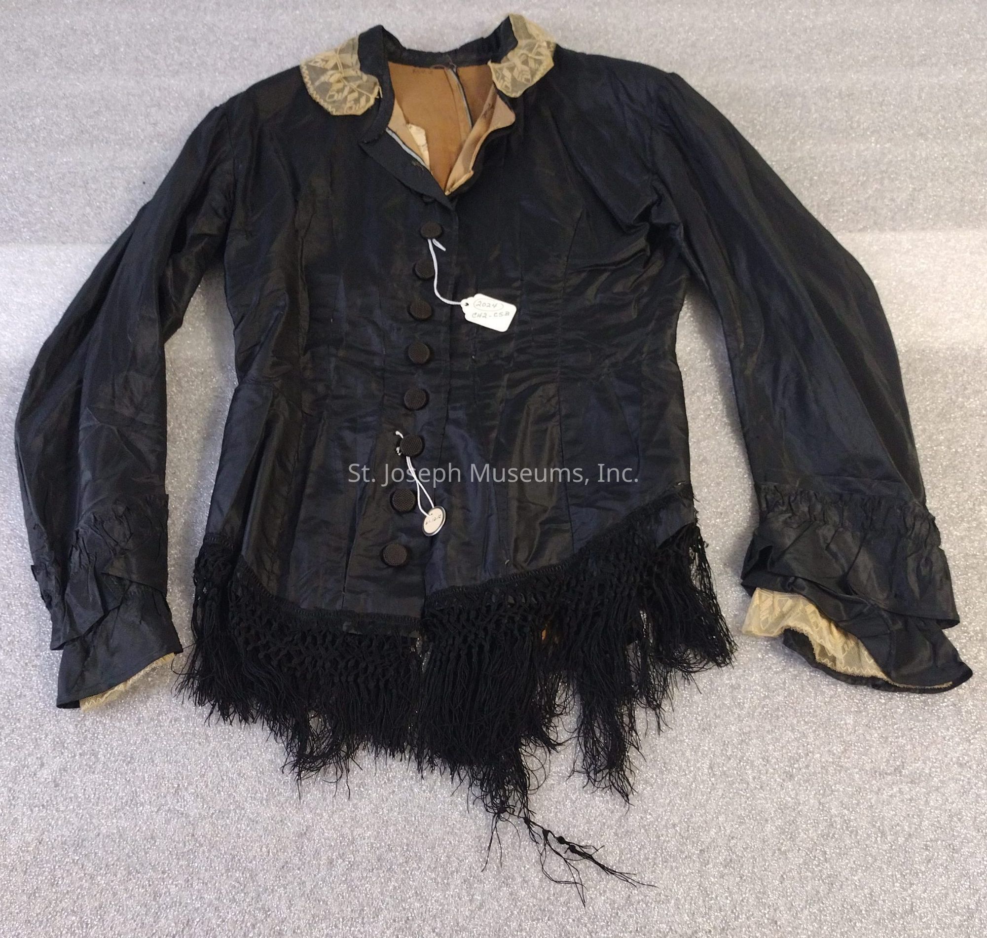 The image depicts a black jacket with a tailored fit, featuring a row of buttons down the front and a distinctive fringe detail along the hem. The garment has long sleeves with ruched cuffs and is complemented by a contrasting beige collar. The inner lining appears to be a light brown fabric. The fabric exhibits a silky texture with slight sheen.