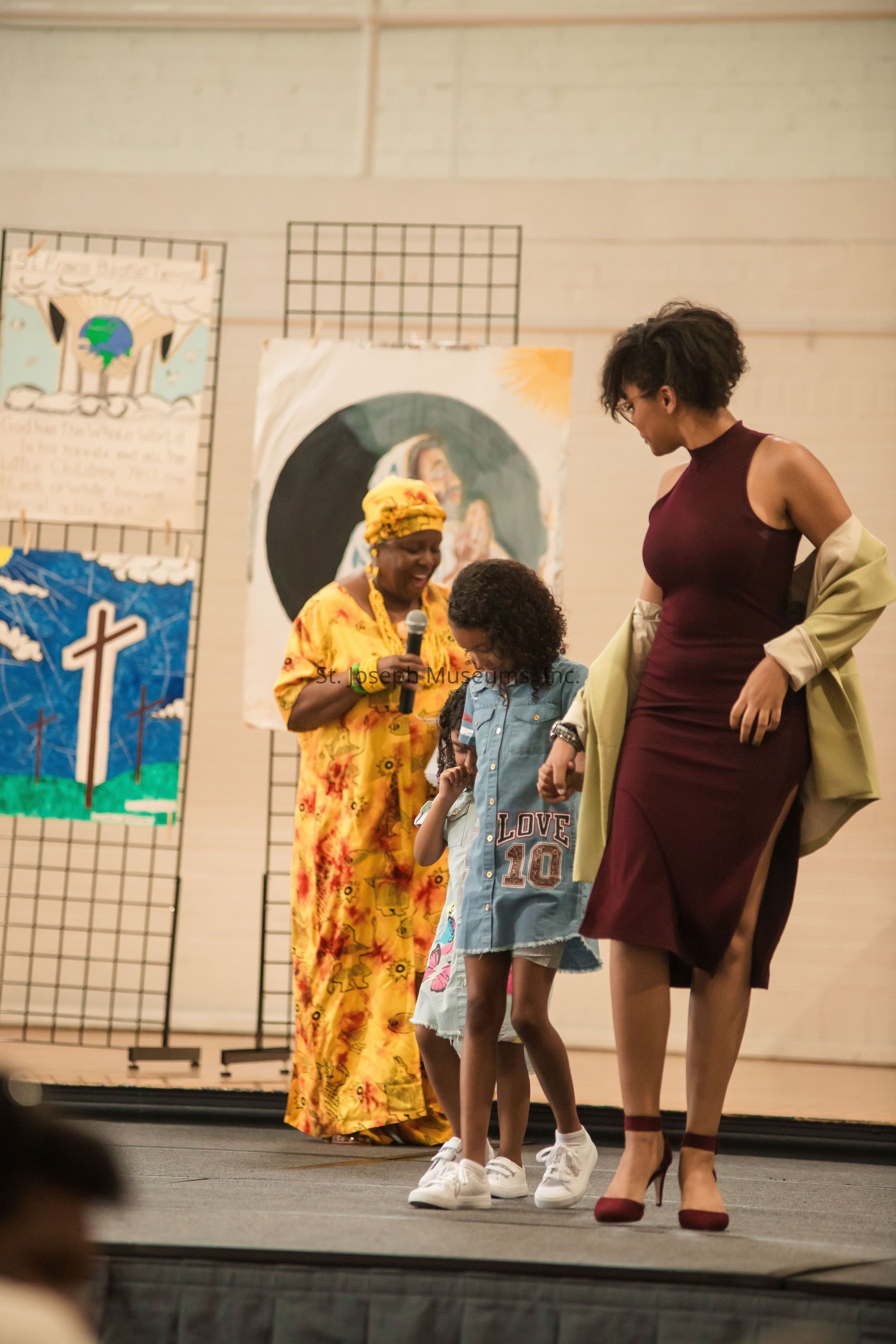 In the image, a woman in a vibrant yellow and orange traditional dress holds a microphone and stands on a stage speaking joyfully to the audience, while next to her, a young girl wearing a denim dress with "Love 10" printed on it is walking alongside another woman in a maroon dress with an elegant cut, who is draping a cream-colored jacket over her shoulders. The setting is a brightly lit indoor venue, and behind them are artistic posters displayed on a grid backdrop, one of which features a cross on a blue and green landscape, and another depicting a figure with a halo. The atmosphere suggests a community or cultural event, as the participants appear engaged and lively, contributing to a scene of warmth and inclusivity.