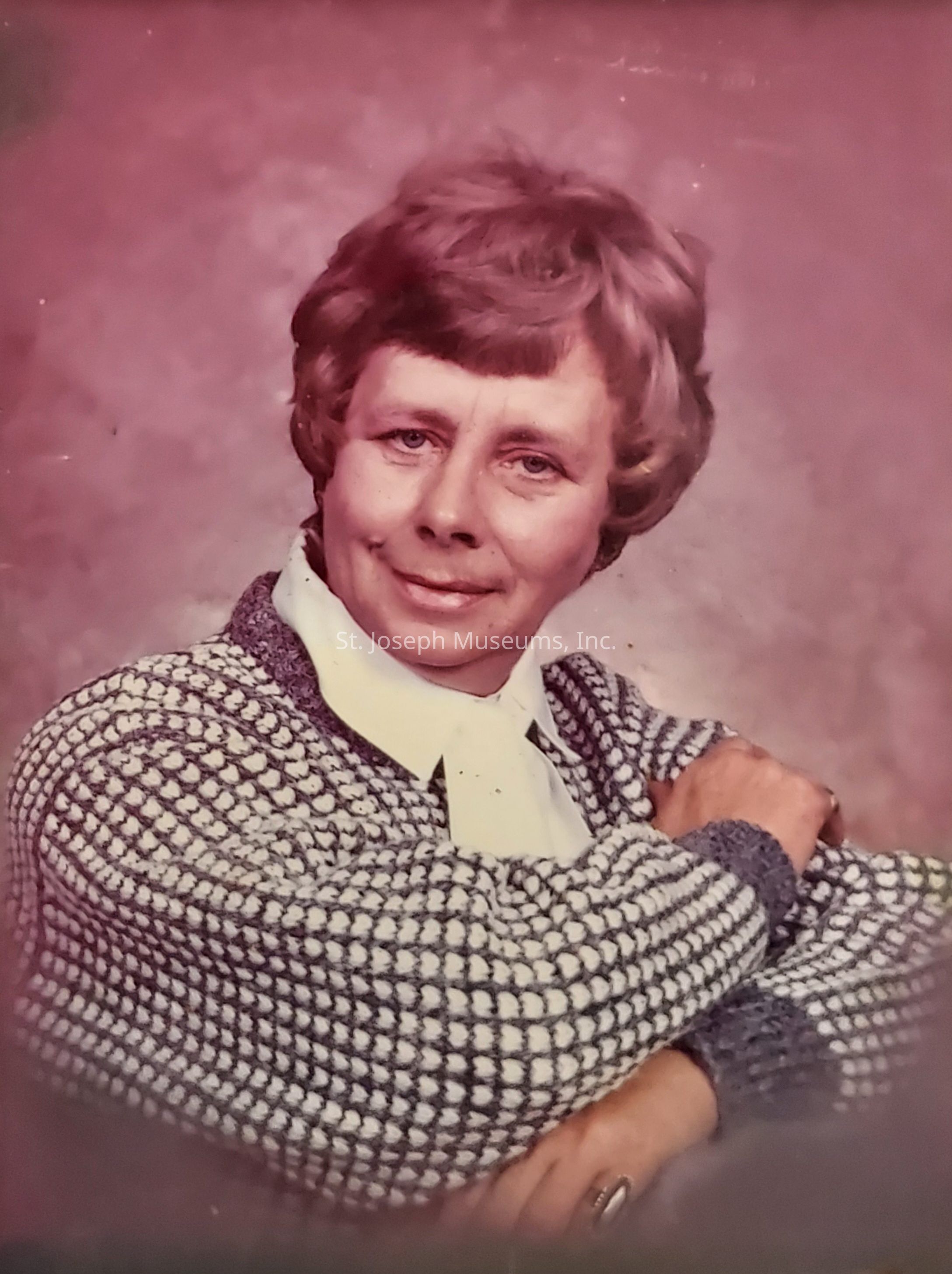 The photograph depicts a middle-aged woman with short, wavy hair, seated in a relaxed pose against a muted, soft-focus background. She is smiling gently, exuding a sense of warmth and approachability. Her attire consists of a textured, patterned sweater featuring a combination of dark and light colors, layered over a crisp white blouse with a neatly folded collar. Her arms are comfortably crossed, suggesting a casual, yet poised demeanor. The overall ambience of the image is nostalgic, capturing a timeless moment that reflects both the style and photographic techniques of its era.
