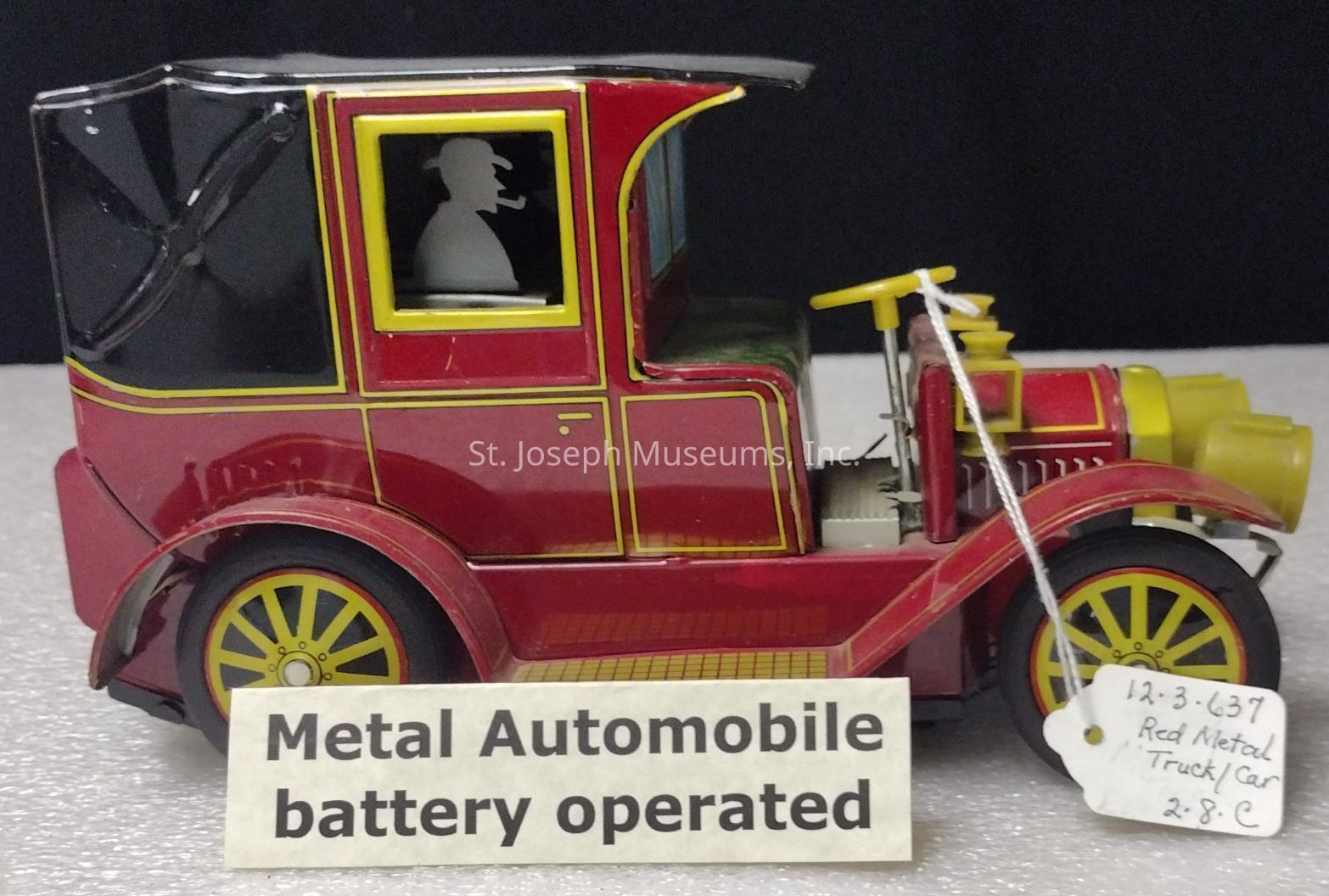 The image features a vintage red and black metal toy automobile that is battery-operated, with a label in front saying "Metal Automobile battery operated." The toy car has yellow accents, including the wheels and steering wheel. A silhouette of a person with a hat and pipe is visible in the window. On the side, a tag attached to the toy reads "12-3-637 Red Metal Truck/Car 2.8-C." The car's design is reminiscent of early 20th-century automobiles, suggesting a classic and collectible item likely intended for display or as a nostalgic piece. The background of the image is plain black, highlighting the car's details.