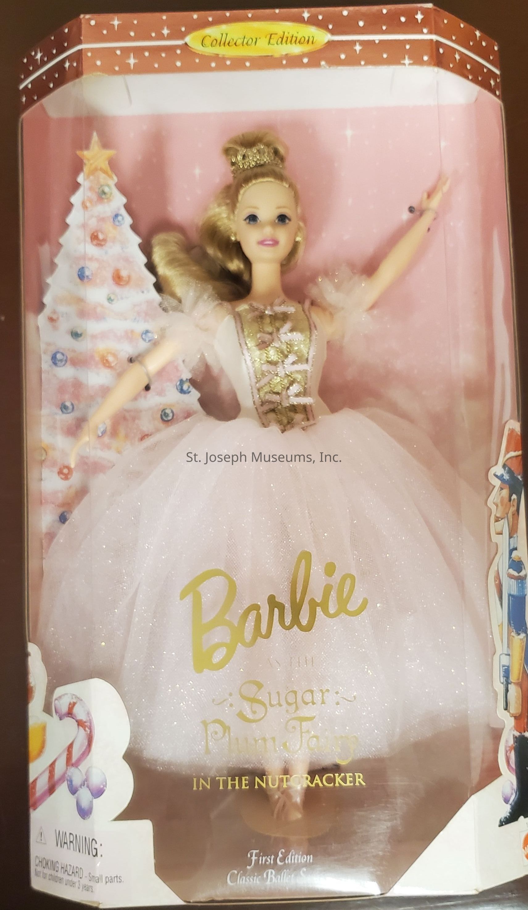The image displays a doll packaged in a box labeled "Collector Edition," showcasing a Barbie dressed as the Sugar Plum Fairy from "The Nutcracker." This first edition of the Classic Ballet Series features Barbie in a shimmering white gown with gold accents, crowned with a tiara matching her elegant ensemble. The packaging includes festive imagery, such as a decorated Christmas tree and a nutcracker, enhancing the seasonal theme. A warning label at the bottom left indicates small parts, cautioning against use by children under three years old. The overall presentation is vibrant and celebratory, appealing to collectors.