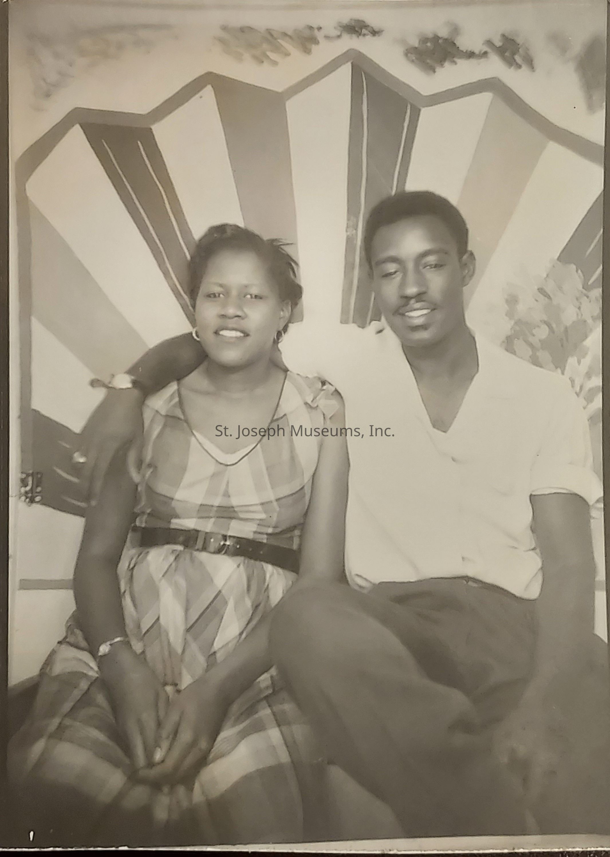 In this black and white photograph, a couple is seated closely together, exuding a sense of warmth and connection. The man, dressed in a light-colored shirt with sleeves rolled up, has a relaxed and content demeanor, while gently placing his arm around the woman beside him. She wears a checkered dress cinched at the waist with a dark belt, and her expression is both serene and inviting. The backdrop features an abstract design with bold lines and patterns, adding a dynamic element to the composition. Each individual's body language indicates comfort and ease in one another's presence, showcasing a moment of shared happiness and companionship.