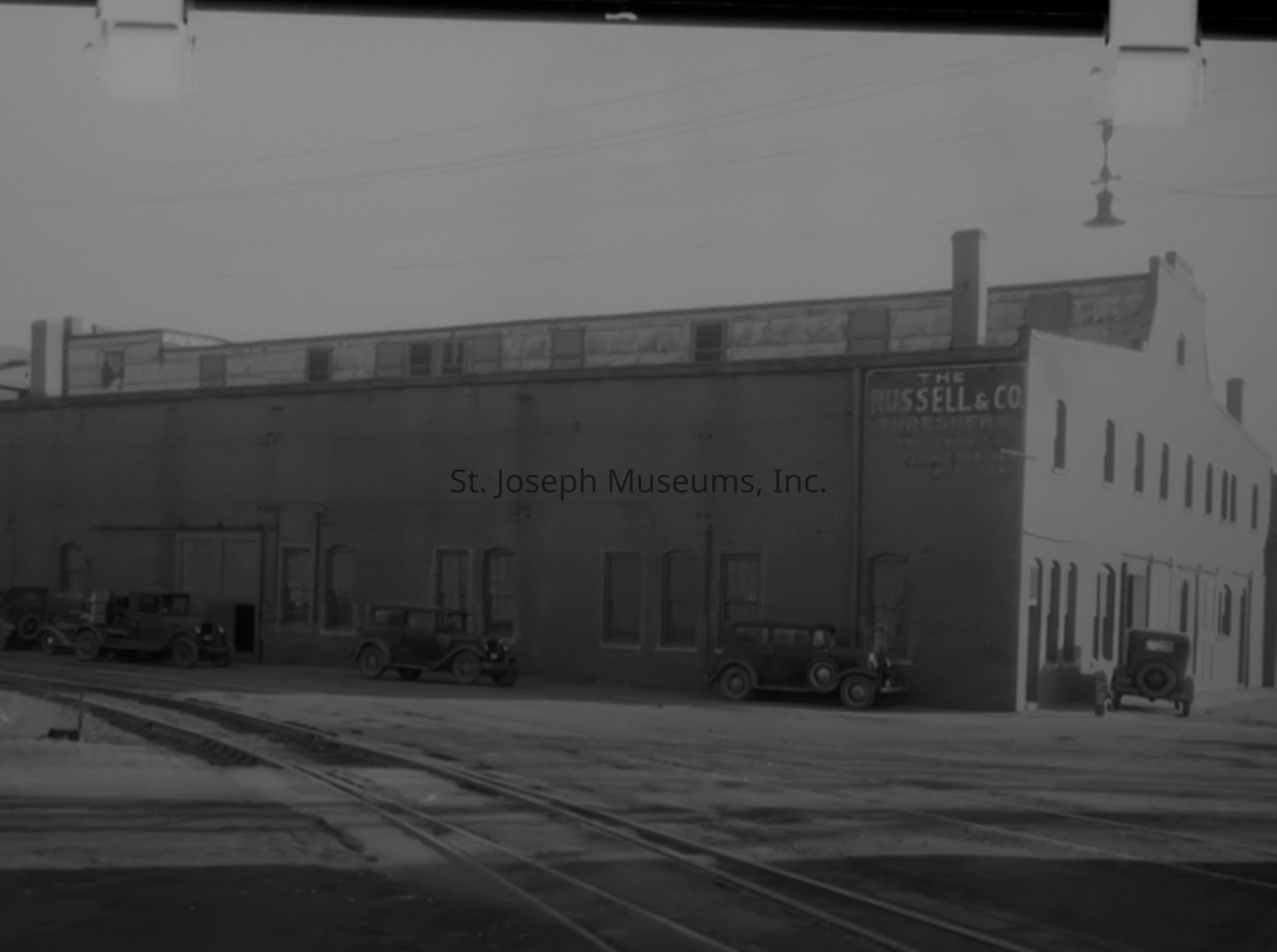 A black-and-white photo depicting a two-story brick building belonging to Russell & Co. Threshers Sales & Service, located at the northeast corner of 5th and Olive Street in St. Joseph, Missouri, from the 1920s; several 1920s-era trucks are parked outside, and a sign is visible on the building's side.