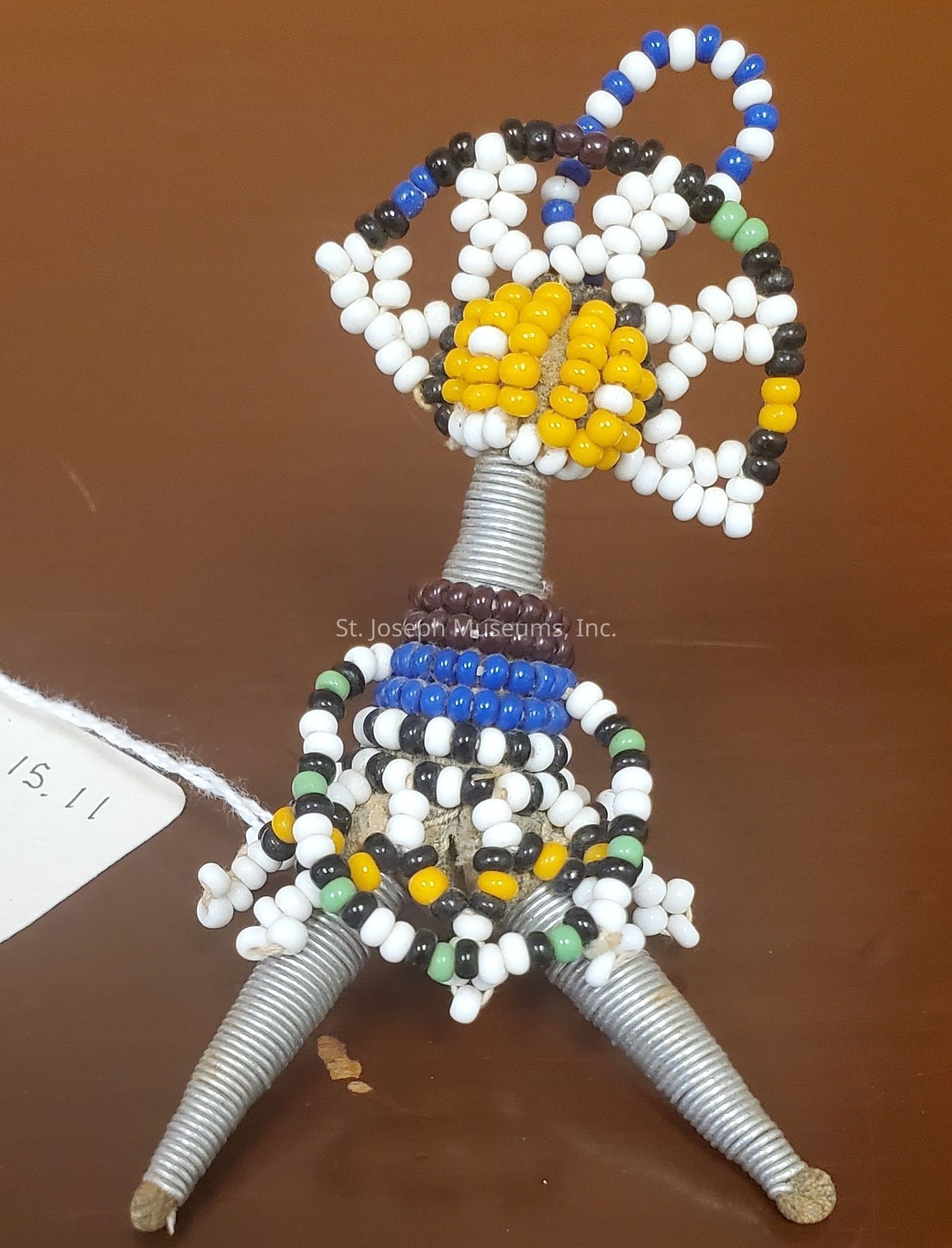 The image depicts a miniature sculpture crafted from colorful beads and coiled wire. The structure features a conical base wrapped in metallic wire, topped by an intricate arrangement of beads in various colors such as black, white, yellow, blue, and green. At the sculpture's apex is a circular formation of beads, resembling a flower or an abstract design, with a prominent yellow central cluster. Additional loops of beads encircle the sculpture, adding layers and texture, while the arranged elements create a vibrant and eye-catching piece that stands out against the plain background.