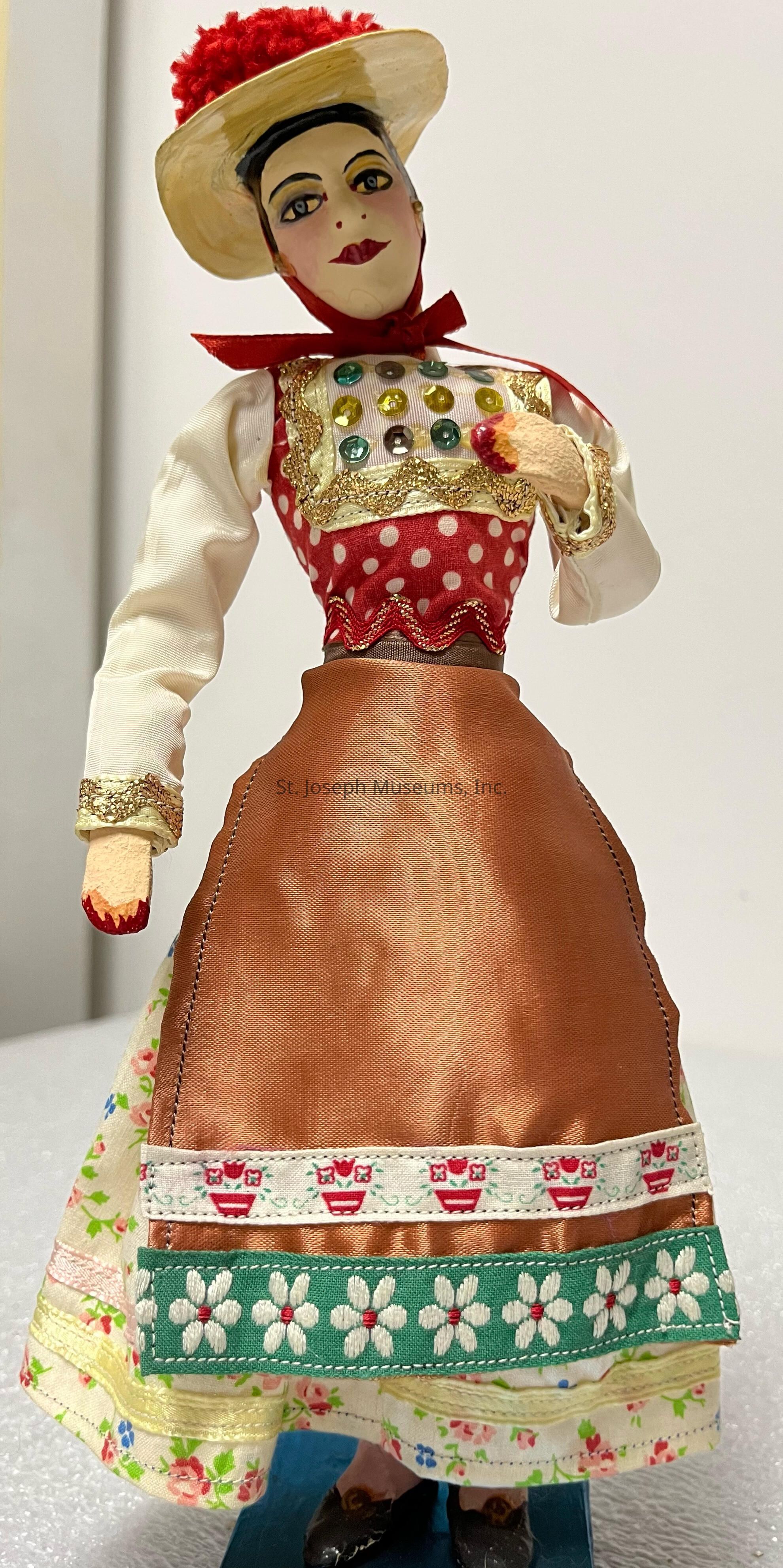 The image features a colorful doll dressed in traditional attire, characterized by a wide-brimmed hat adorned with red embellishments and a red scarf around the neck. The doll wears a polka-dotted top with decorative sequins and gold trim, and its sleeves are long and white with gold lace at the cuffs. The lower part consists of a copper-colored apron layered over a floral-print dress with intricate embroidery at the hem, showcasing patterns of flowers and small, red cup-like designs. The doll stands on a blue platform, adding a sense of stability and highlighting its intricate craftsmanship and vibrant cultural representation.