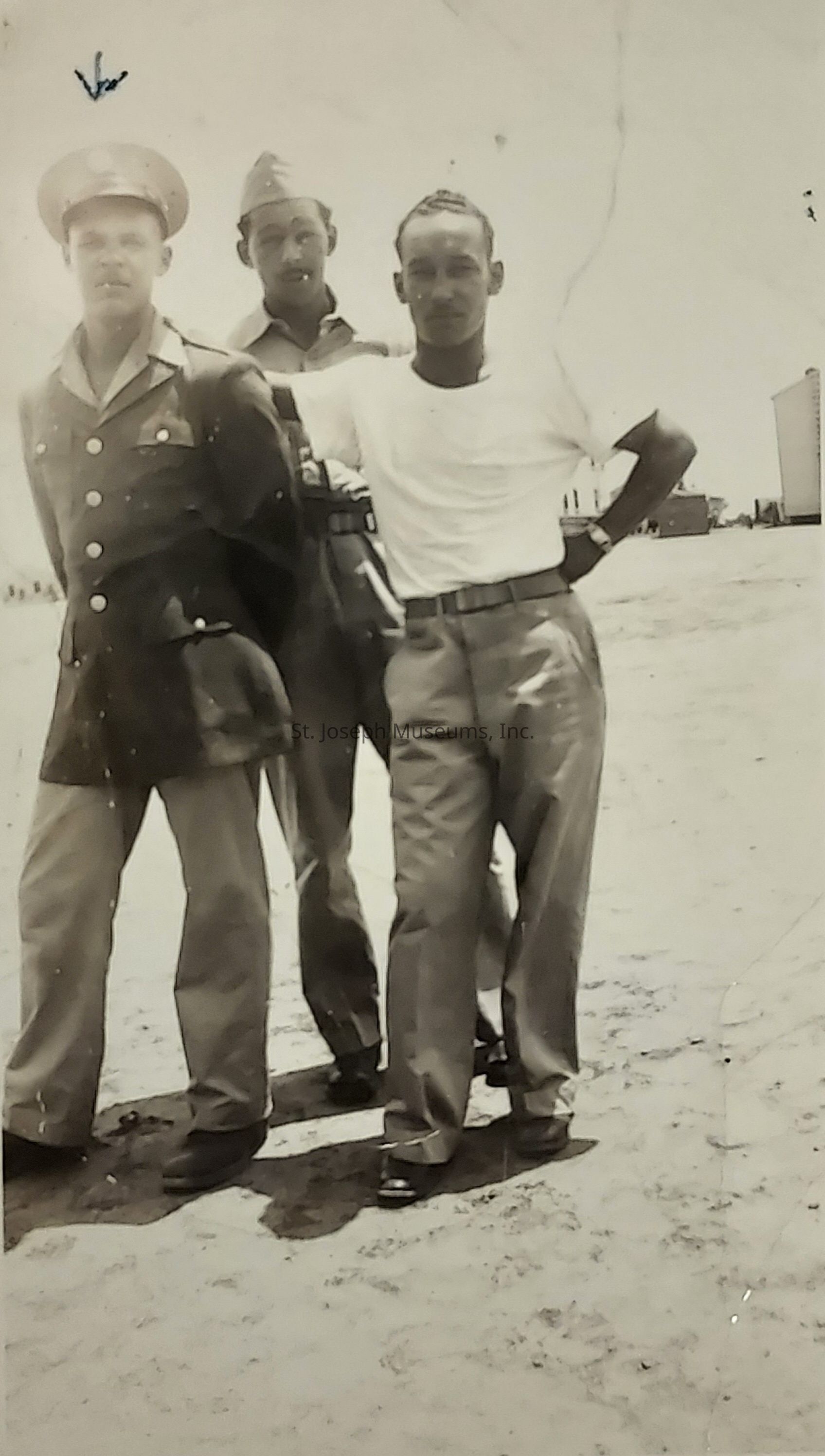 The image depicts three men standing together in a vintage photograph, likely from a mid-20th century military setting, given their attire. The man on the left is wearing a military uniform with a cap, complete with insignia, while the man in the middle wears another uniform with a soft cap. The man on the right is dressed in a casual, plain white t-shirt with his hands on his hips, projecting a relaxed demeanor. They are standing on an open terrain, possibly a military base, with buildings visible in the distant background. The photo is black and white, capturing a moment in what seems like bright daylight, evidenced by the shadows cast on the ground. The men's expressions are serious yet relaxed, suggesting camaraderie or a break during their duties. The overall setting and the attire of the men evoke a sense of historical context, possibly during a period of training or deployment.