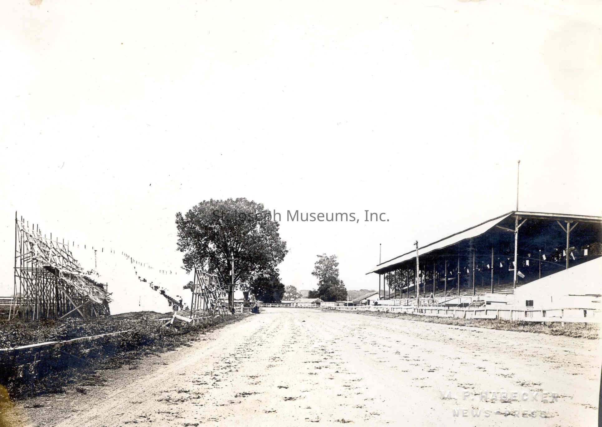Pony Express Pageant Stage construct at Lake Contrary, 1923: Black-and-white photograph from August 8, 1923, depicting the construction of a 600-foot stage at Lake Contrary Driving Park, where workers are setting up the platform on the left side to host the Pony Express pageant scheduled to recreate the development of St. Joseph from 1843 to 1860. A covered grandstand is visible in the distance on the right, while a dirt track stretches through the foreground, capturing the scene's eastward view in preparation for the historical spectacle.