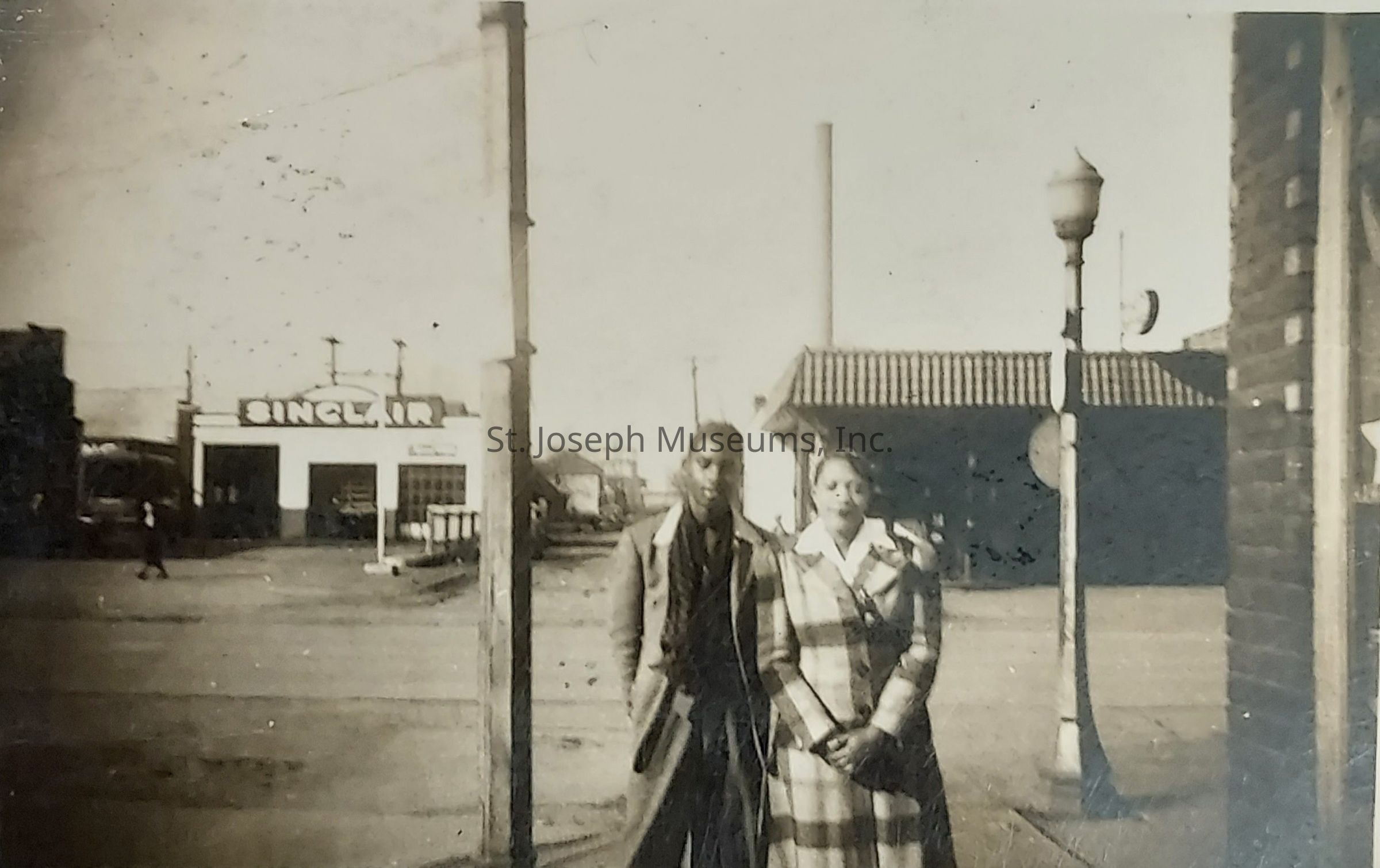 The black and white photograph captures a vintage street scene with a man and a woman standing side by side in the foreground. The man is dressed in a long coat, and the woman wears a plaid coat. They are positioned near what appears to be a street lamp and a wooden post. Behind them, there is a building with a sign that reads "SINCLAIR," indicating it may be a gas station or garage. The background features additional structures, including a building with a tiled roof and a chimney. The image conveys a sense of history, with architectural elements and fashion reflective of an earlier era.