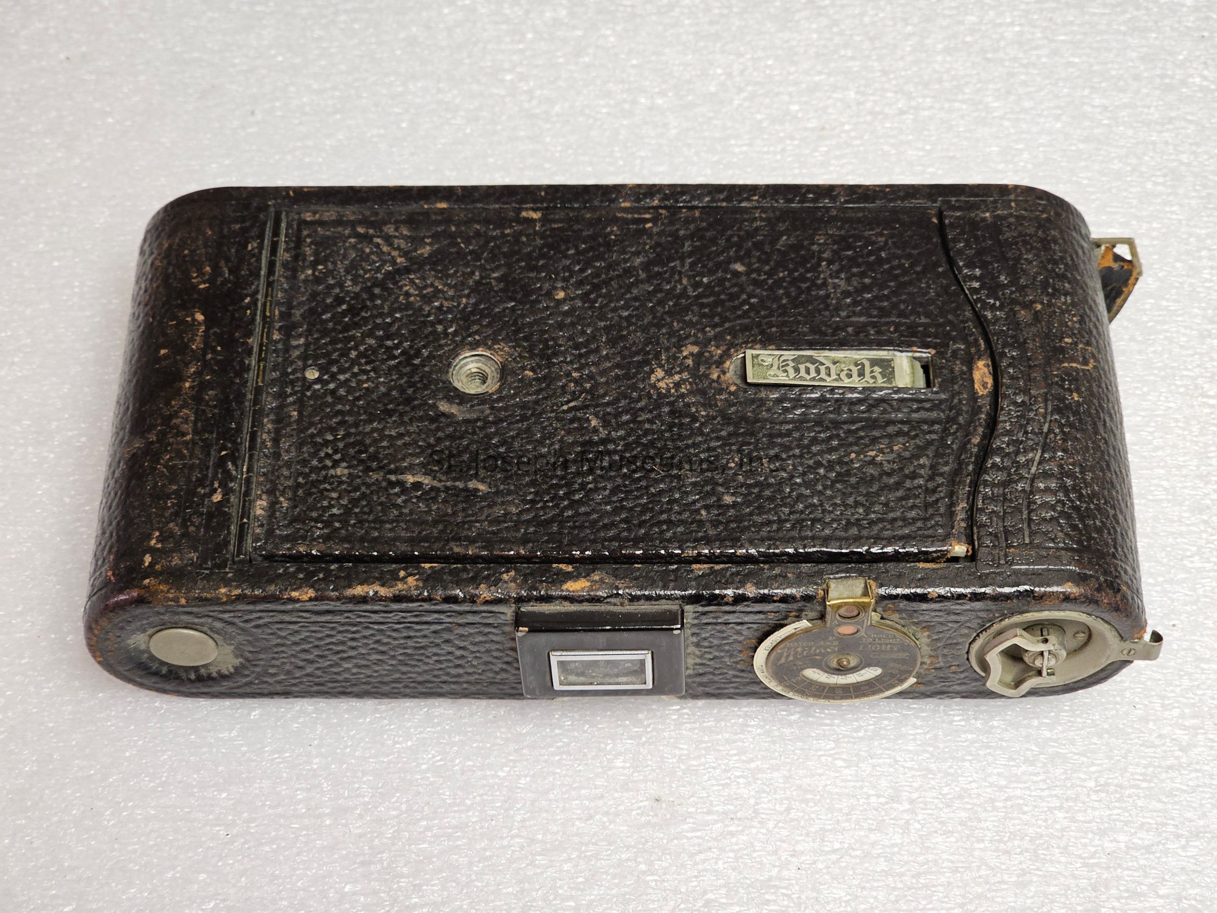 The image shows an antique folding camera with a worn, textured black leather exterior and metal accents displaying age-related patina, indicating its vintage nature. On the front panel, the recognizable "Kodak" nameplate is prominently displayed, suggesting its manufacture by the iconic camera company. The camera's body, which appears to be made of sturdy materials, features various mechanical components such as knobs and a winding mechanism, hinting at the manual operations required during its use. There is also a circular metal dial on the right side beside the winder, possibly used for adjusting settings. The overall craftsmanship, marked by simplicity yet effective design, points to a time when photography was both an art and science, requiring knowledge and skill. This object evokes a sense of nostalgia, reminiscent of early 20th-century photography practices, capturing history through the lens of a technological relic.