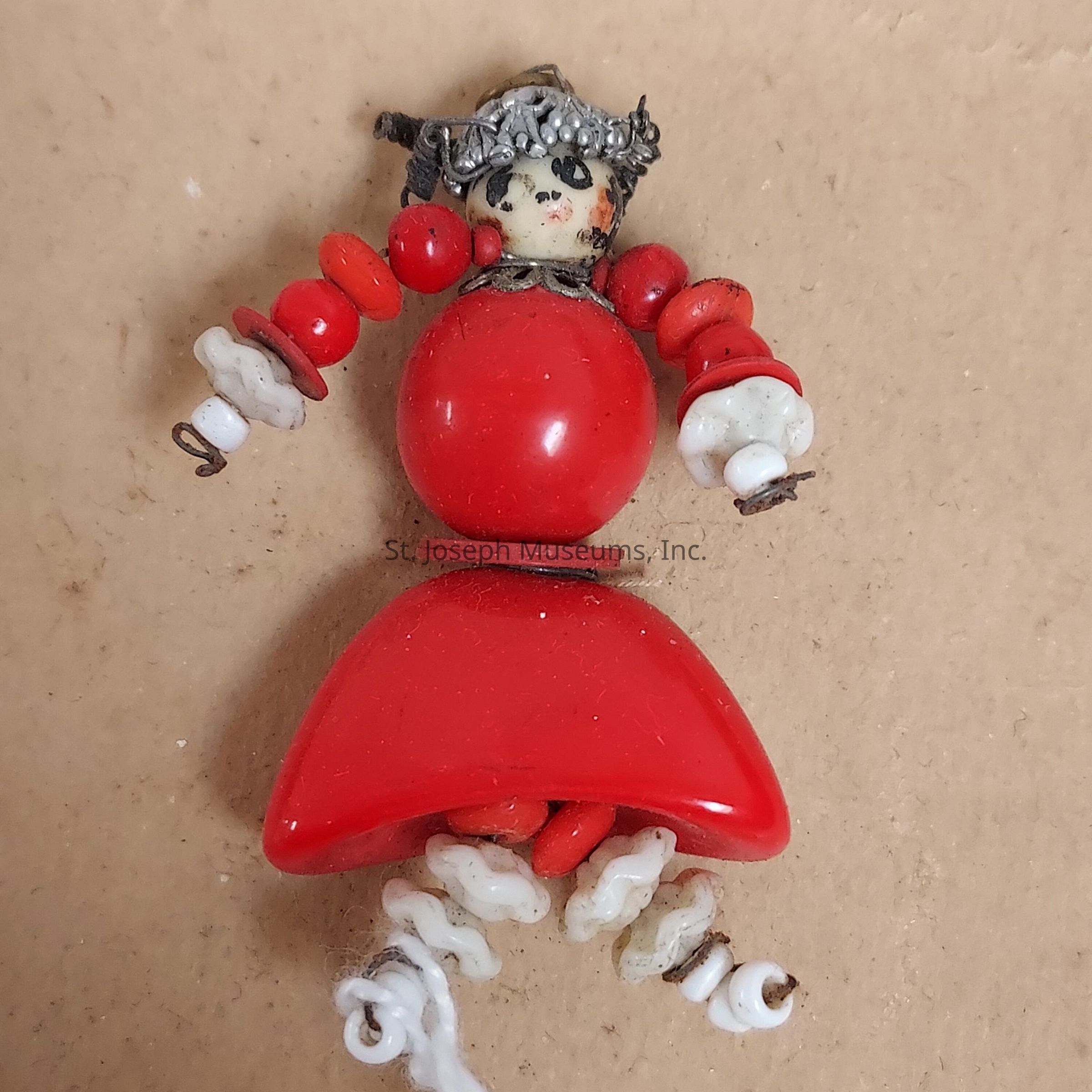 The image shows a handmade doll-like figure composed of various colored beads. The figure has a head made of a small, painted bead that includes facial details with black dots for eyes and other slightly faded features, along with a metallic headpiece. The body consists of larger red beads, forming a round upper section and a skirt-like lower section. The arms are articulated with red and white beads, and the hands have small loops at the ends. The legs are made of intertwined white beads, resembling a spiral or twisted design, and terminate in similar loops as the hands, suggesting points for attachment or movement. The entire assembly is laid on a light brown surface, which provides a plain contrast to the vibrant red of the beads, highlighting the artistic and handcrafted essence of the figure. This unique and creative piece represents a fusion of craft and design, showcasing the imaginative use of simple materials to create a distinct three-dimensional object that evokes a sense of whimsy and charm.