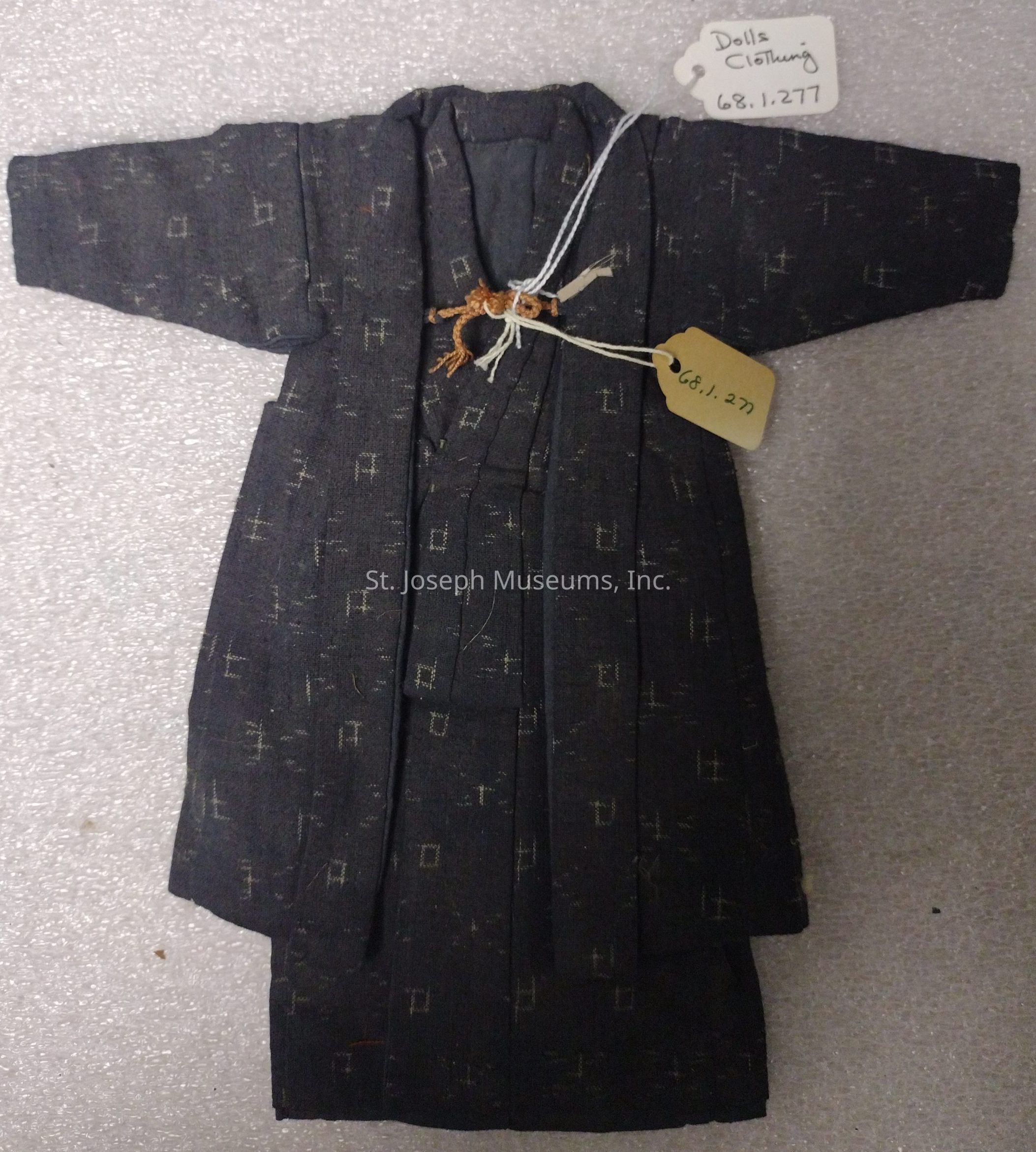 The image shows a small, traditional-style garment for a doll, resembling a kimono, made from a dark fabric with a subtle pattern of small square shapes. The garment is laid flat on a textured surface, displaying its wide sleeves and layered look typical of kimono-like clothing. There are two tags attached to the garment: one of them, white with black writing, reads "Dolls Clothing 68.1.277," while the other, a smaller beige tag, also displays the number "68.1.277." These tags are secured with strings, hinting that this piece might be a cataloged item, possibly in a museum or a collection. The intricate design and careful craftsmanship of the garment suggest attention to detail and an emphasis on maintaining cultural elements even in miniature form, reflecting the aesthetic values often associated with traditional clothing. The fabric texture and the shadows add depth to the visual presentation, emphasizing the garment's material and style. This depiction captures a blend of cultural heritage and artistry preserved