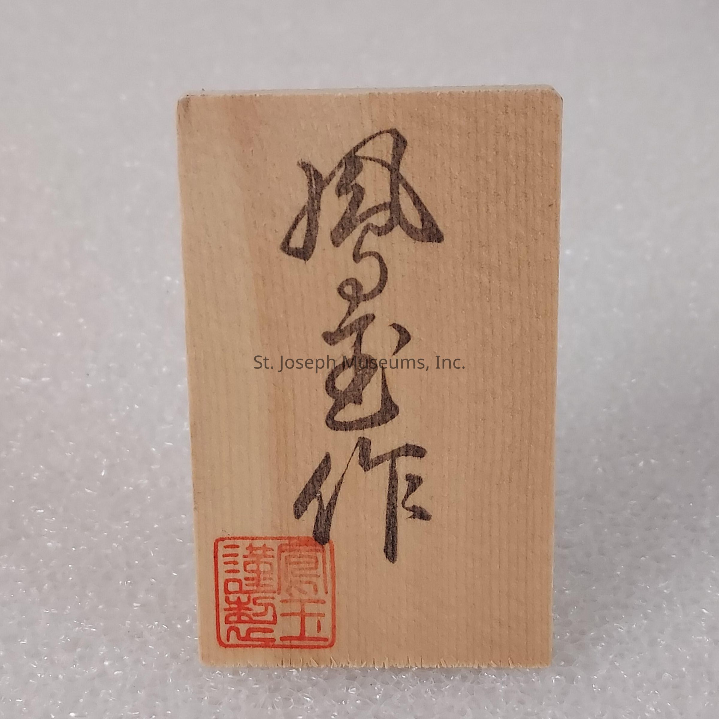 The image shows a small wooden plaque featuring Japanese calligraphy, with a vertical line of characters starting from the top. The wood has a natural grain, and the calligraphy appears to be carefully hand-painted in a dark ink. At the bottom, there is a red ink stamp, which might be a seal or signature, indicating authenticity or artistic identity. The background consists of a textured, light-colored surface, which contrasts with the smooth appearance of the wooden plaque, effectively highlighting the craftsmanship and traditional art style.