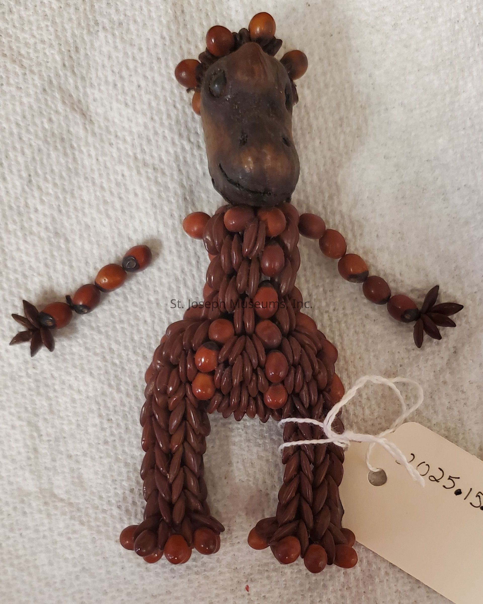 The image shows a whimsical, handmade figurine shaped like a horse, crafted entirely from brown seeds and pods. The head has a textured surface with small, dark bead-like eyes, and a subtle, rounded snout. The mane is formed by a series of round, smooth seeds that encircle the top of the head. The main body and legs are constructed with elongated, darker seeds arranged closely together, creating a braided appearance. The arms are made from a series of interconnected round seeds, ending in star-shaped seed "hands." The entire figure rests on a plain, white fabric background, with a small, white tag attached to one leg, bearing the numbers "2025.153" written on it.