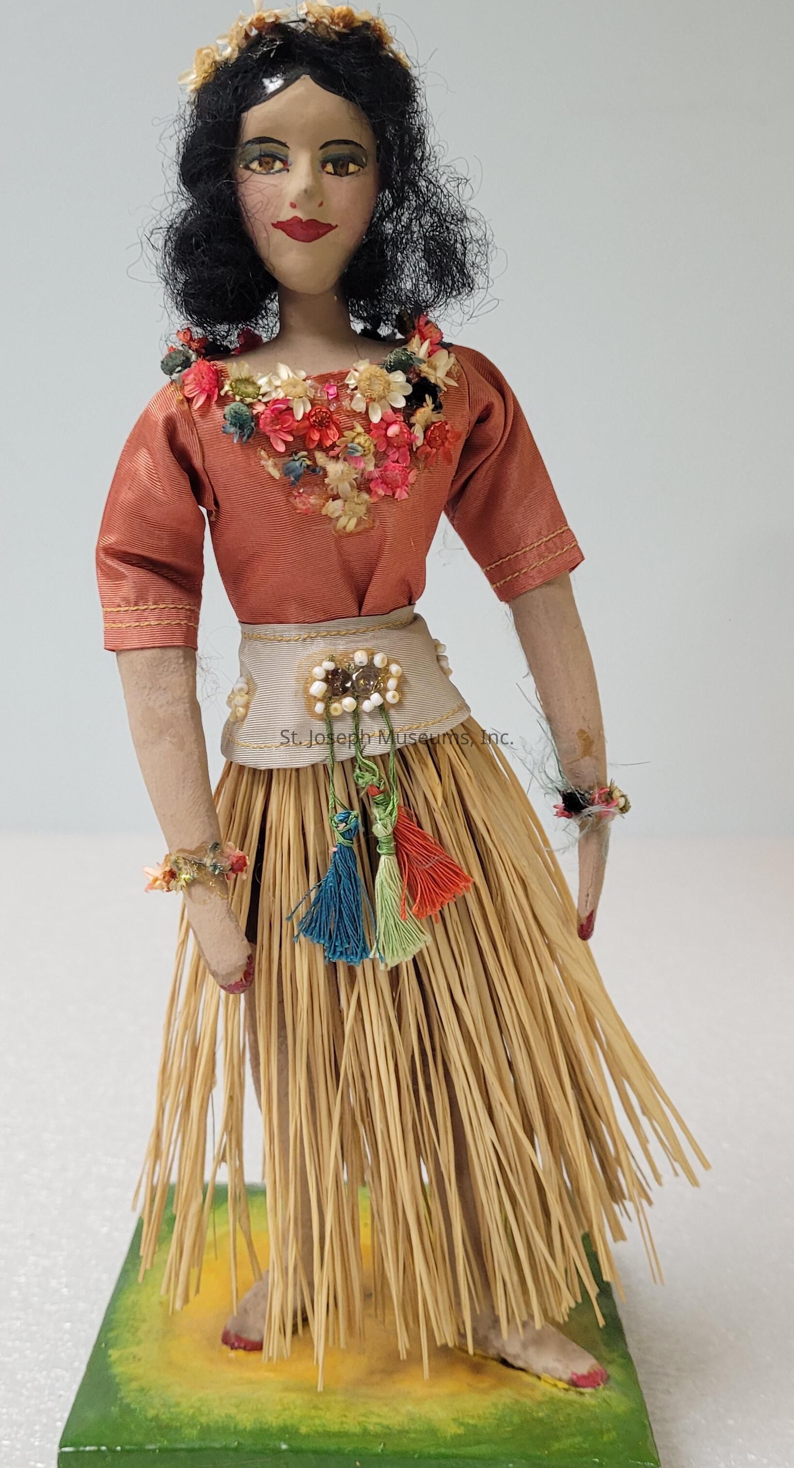 The image depicts a handcrafted doll adorned with detailed elements that contribute to its cultural representation and artistic charm. The doll stands on a painted green and yellow base, showcasing a face with striking, painted features, including red lips and expressive eyes. It wears a coral-colored blouse and a floral necklace, adding vibrant pops of color to its attire. Above the waist, a white fabric band embellished with beads and tassels in blue, green, and red catches the eye. The doll's skirt is made of a natural material resembling straw, reminiscent of traditional hula costumes, emphasizing its cultural theme. A crown of flowers adorns the doll's dark hair, further enhancing its picturesque and festive appearance. The meticulous detailing in the floral patterns, the careful arrangement of tassels, and the texture of the materials all contribute to a rich presentation, showcasing the artisan's skill. The surrounding colors and textures complement each other, providing a charming and lively aesthetic.