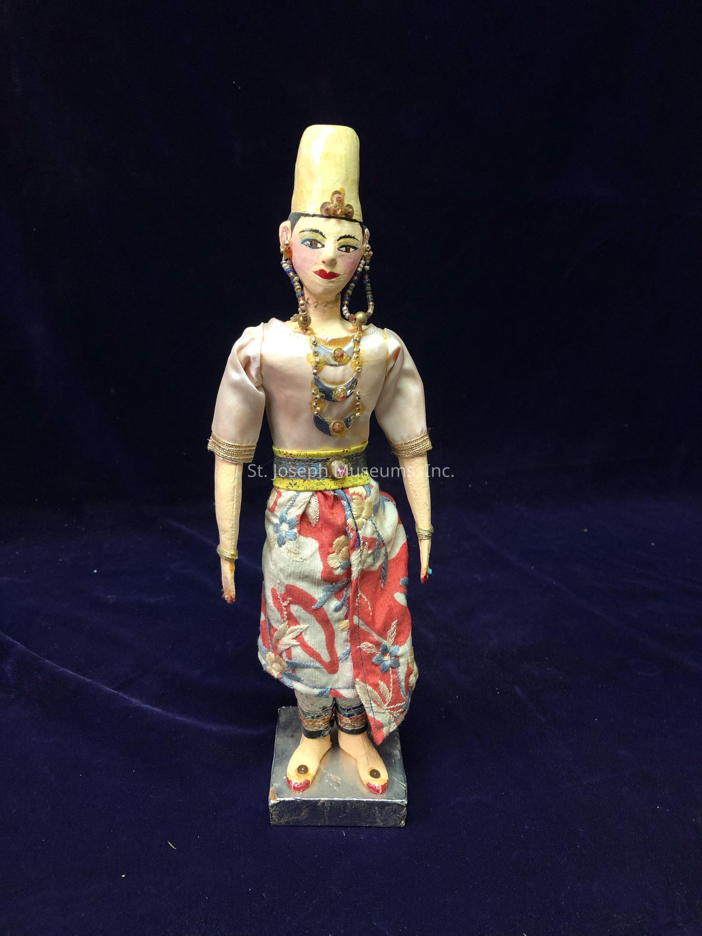 The image depicts a handcrafted doll standing upright against a dark, textured background. The doll wears an ornate beige blouse paired with colorful, floral-patterned pants, exuding an air of cultural richness. Its accessories include elaborate necklaces, and the head is topped with a tall, sculpted golden hat, enhancing its royal appearance. The facial features are meticulously painted, with striking eyes, rosy cheeks, and red lips, adding expressive character. Further detailing can be seen in the earrings and arm bands, which add to the overall intricate design. The doll stands on a rectangular base, which provides support and highlights its stately pose.