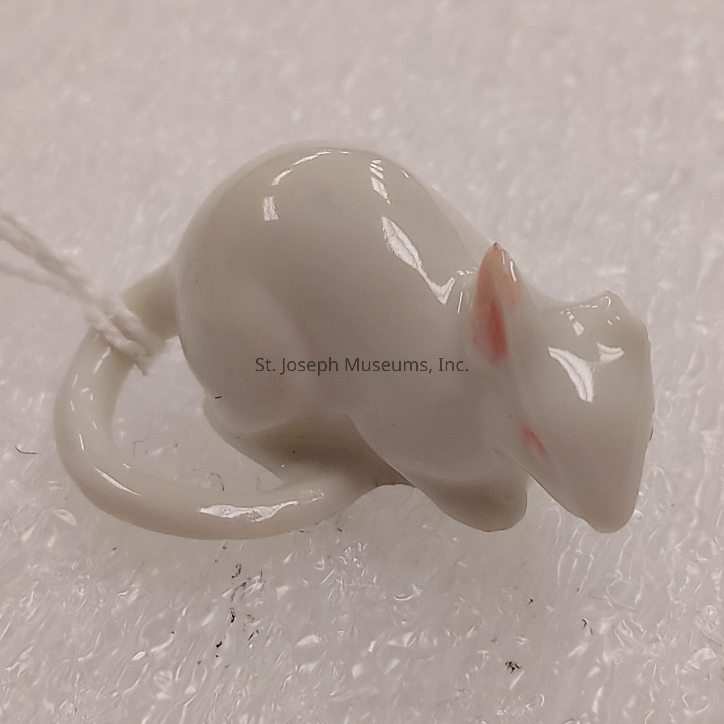 The image shows a shiny, small porcelain figurine of a mouse on a textured white surface. The mouse is depicted with a slightly hunched posture, its tail curling around its body. The surface has a glossy finish, allowing light reflections that emphasize the smoothness of the porcelain material. Slightly visible are its pink-tinged ears and delicate features, which add subtle detail to the figure. Attached to the figurine’s tail is a string, possibly for hanging purposes, as it could be an ornament. The background further highlights the figure’s sheen due to its contrasting mat appearance.