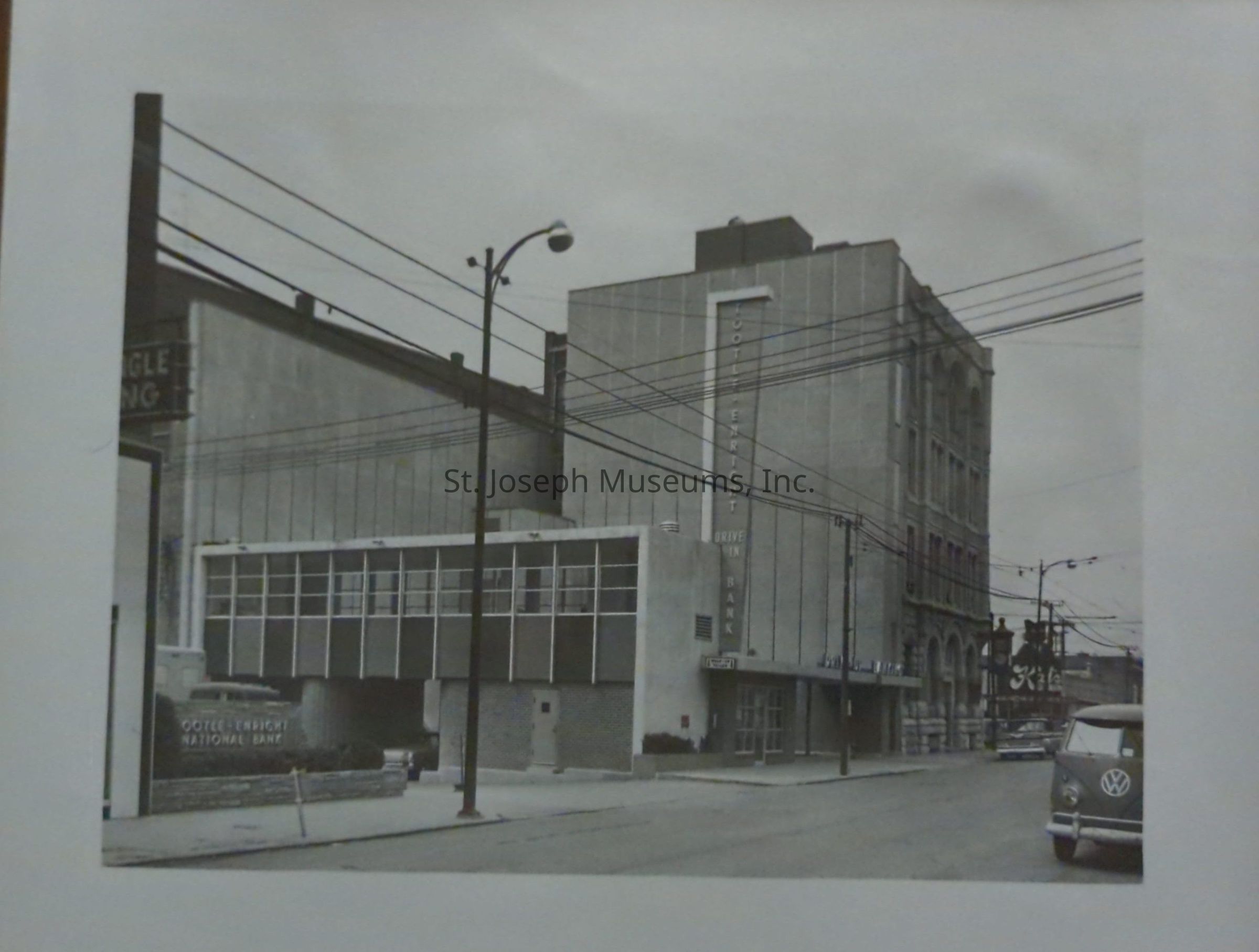 Black-and-white image showcasing Edmond Street looking west toward 6th Street, featuring the Tootle-Enright Bank. In the foreground, there is a Volkswagen van parked on the street, with several utility lines crossing above. The architecture of the bank is prominent, characterized by its mid-20th-century design, and there are additional buildings visible further down the road.