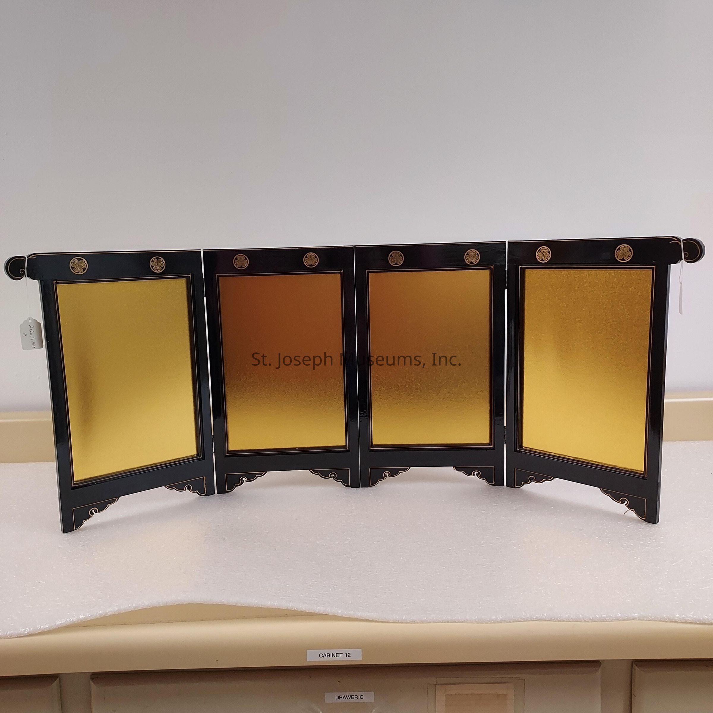 The image depicts a decorative folding screen with four panels, each featuring a vibrant gold finish framed by a sleek, black border. The top of each panel is adorned with small, circular accents, which appear to be ornamental details adding elegance to the design. The screen is set against a plain, light-colored background, highlighting its rich, gold tone. The panels are arranged in a slightly curved manner, suggesting flexibility and adaptability to different settings. Positioned atop a surface that resembles light-colored wood or a similar material, the screen can be utilized as a sophisticated backdrop or room divider, combining aesthetic appeal with functional versatility. The tag attached on the left suggests it might be part of an exhibit or for sale.