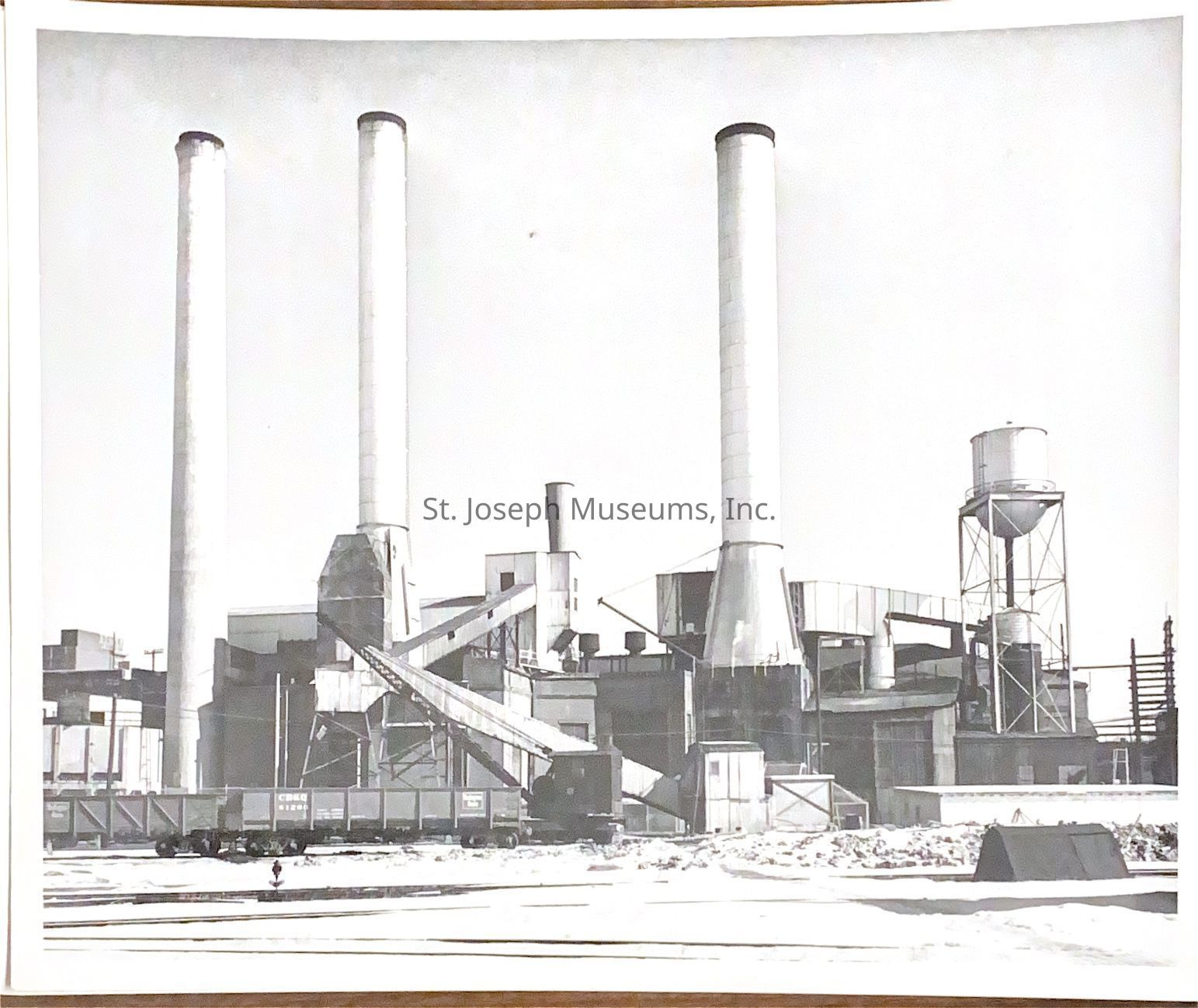 Black-and-white photograph of four tall smokestacks of the St. Joseph Light and Power Company at the intersection of Felix and Main Streets, surrounded by industrial structures, including a conveyor belt system and a water tower, creating a skyline dominated by industrial architecture and machinery.
