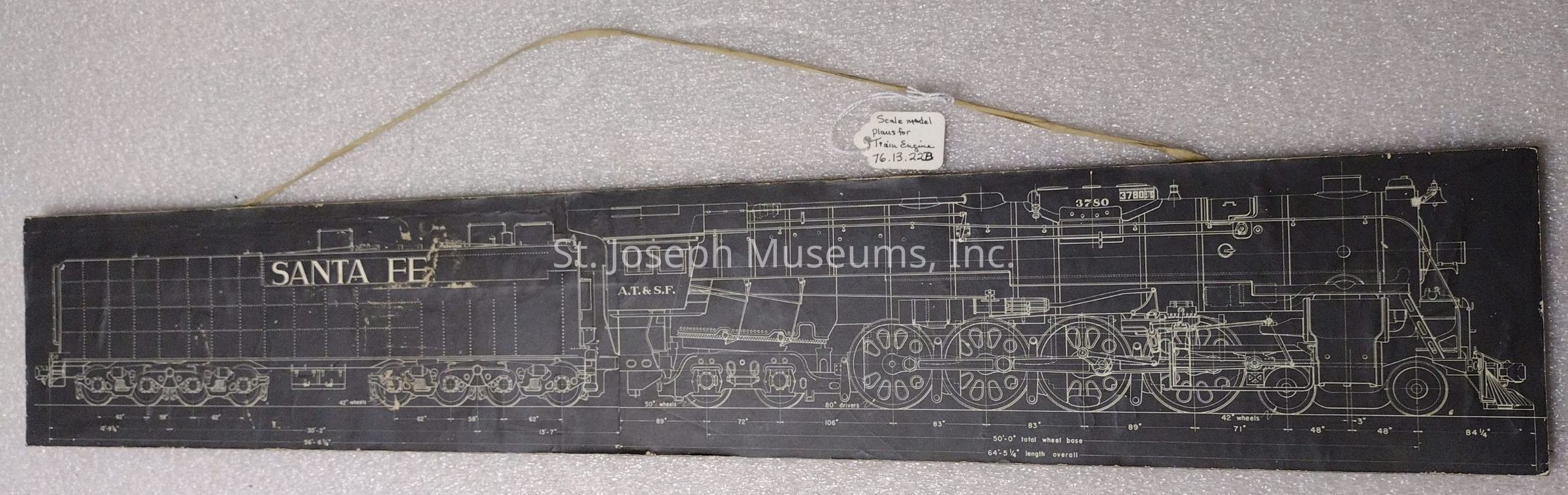 The image depicts a detailed blueprint drawing of a steam locomotive, labeled with "Santa Fe" on the tender and "A.T. & S.F." on the engine, showcasing intricate technical details such as the wheels, axles, and various components of the locomotive structure, all rendered in a white line drawing on a black background. Measurements and scale indicators are clearly marked, providing precise dimensions and specifications that imply the document's purpose as a scale model plan. A small tag attached to the top of the plan reads "Scale model plans for Train Engine 76.13.22B," indicating it is a reference material or historical artifact, possibly used for educational or restoration purposes, and the entire layout is designed to convey the complexity and sophistication of early 20th-century locomotive engineering. The drawing is presented on a textured, foam-like surface that contrasts with the smoothness of the plan itself, suggesting a display or archival storage setting intended for preservation and study.