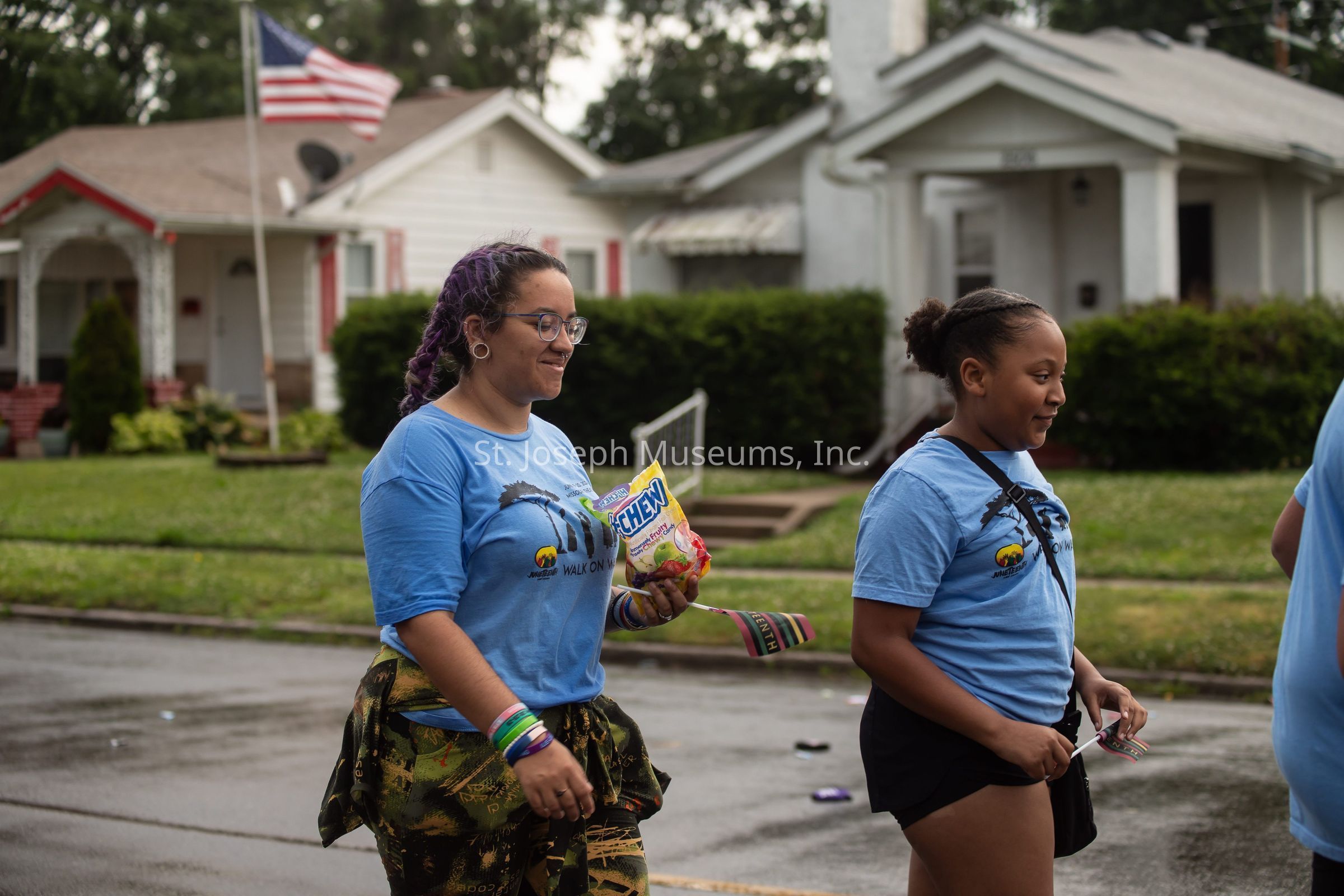 In the image, two people are walking together on a wet street in what appears to be a residential neighborhood. Both are wearing blue T-shirts, indicating they might be part of an event or organization. The person on the left has long purple hair and glasses and holds a bag of Hi-Chew candy, while also sporting colorful wristbands. The person on the right is younger, with their hair tied back, and carries some small items in hand. The background showcases tidy lawns and houses with an American flag visible on a flagpole, suggesting a peaceful community setting.