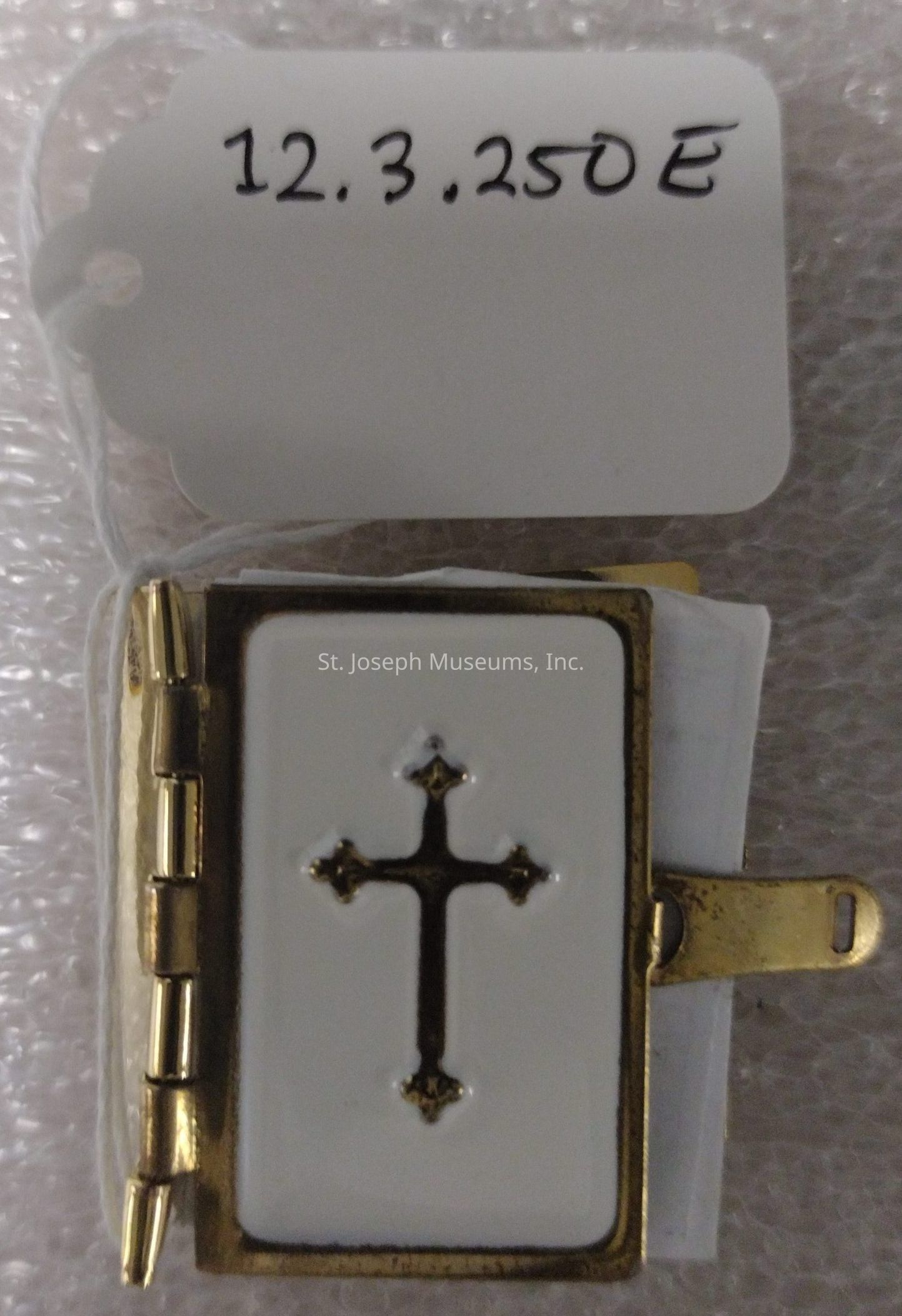 The image shows a small, rectangular, gold-edged book with a white cover featuring an ornate gold cross in the center, attached to a white tag with a string. The tag displays the numbers and letters "12.3.250E" written in black ink. The book appears to have a hinge and clasp mechanism, indicating it might be a decorative item designed to resemble a miniature book or locket, possibly used for religious purposes or as a collectible. The background consists of a textured material, resembling bubble wrap, suggesting the item may be carefully packaged for protection or display. The cross design on the cover has intricate detailing at each end, giving it a traditional and elegant appearance.