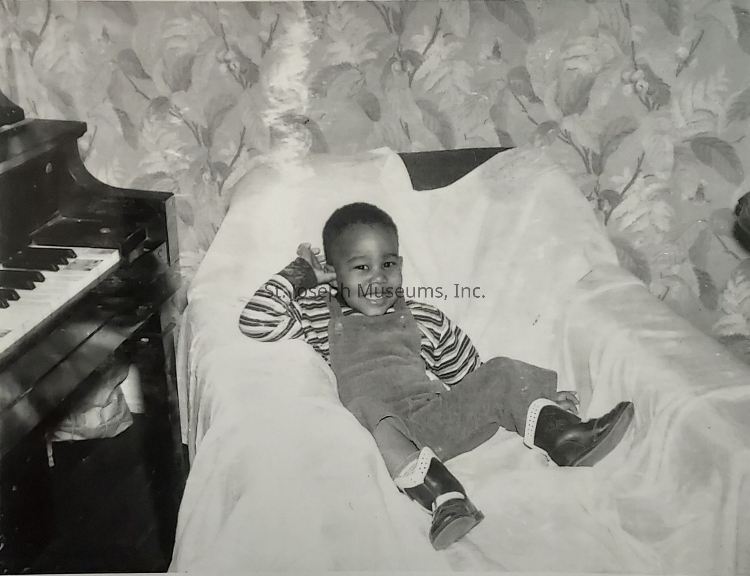 In this black-and-white photograph, a young child is comfortably lounging on a plush armchair, their feet propped up and a hand resting casually behind their head. Clad in denim overalls and a striped shirt, the child sports a cheerful smile, exuding an aura of joy and innocence. To the left of the image, a piano with visible keys stands against a wall adorned with a floral-patterned wallpaper, adding a vintage charm to the setting. The room’s ambiance is cozy and warm, suggesting a home filled with love and music, as evidenced by the presence of the piano, hinting at a possible fondness for musical melodies or family gatherings centered around music. The child’s boots are slightly elevated, adding to the relaxed and playful demeanor captured in this candid moment. The overall scene paints a nostalgic picture, evoking a sense of warmth and happiness, encapsulating a snapshot of carefree childhood days spent in simple comfort and bliss at home.