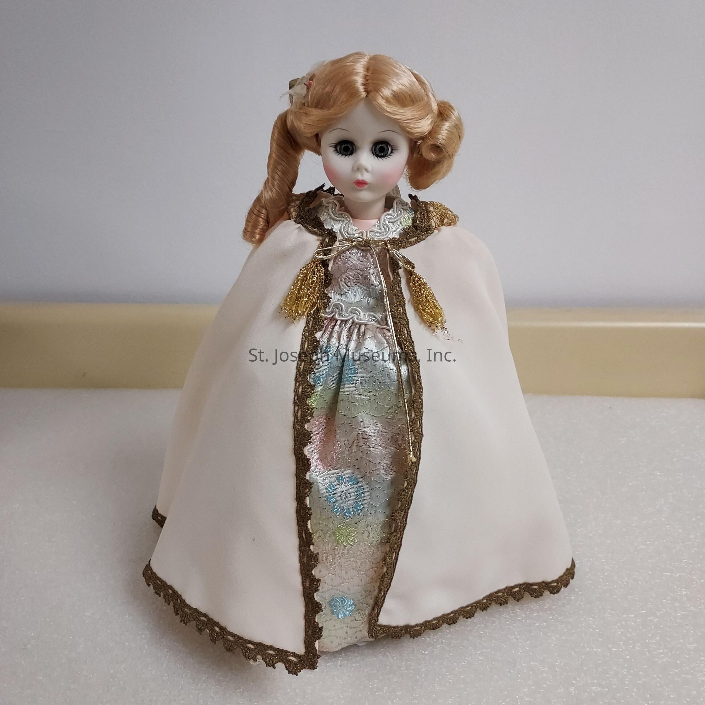 The image shows a porcelain doll with large eyes, styled with curled blonde hair. She is dressed in an elegant, flowing white cape with gold trim along the edges and a matching clasp at the neckline. Underneath, she wears a silvery dress adorned with subtle patterns and hints of pastel colors like blue and pink. The doll is in the likeness of Martha Johnson Patterson, First Lady and daughter of president Andrew Johnson.