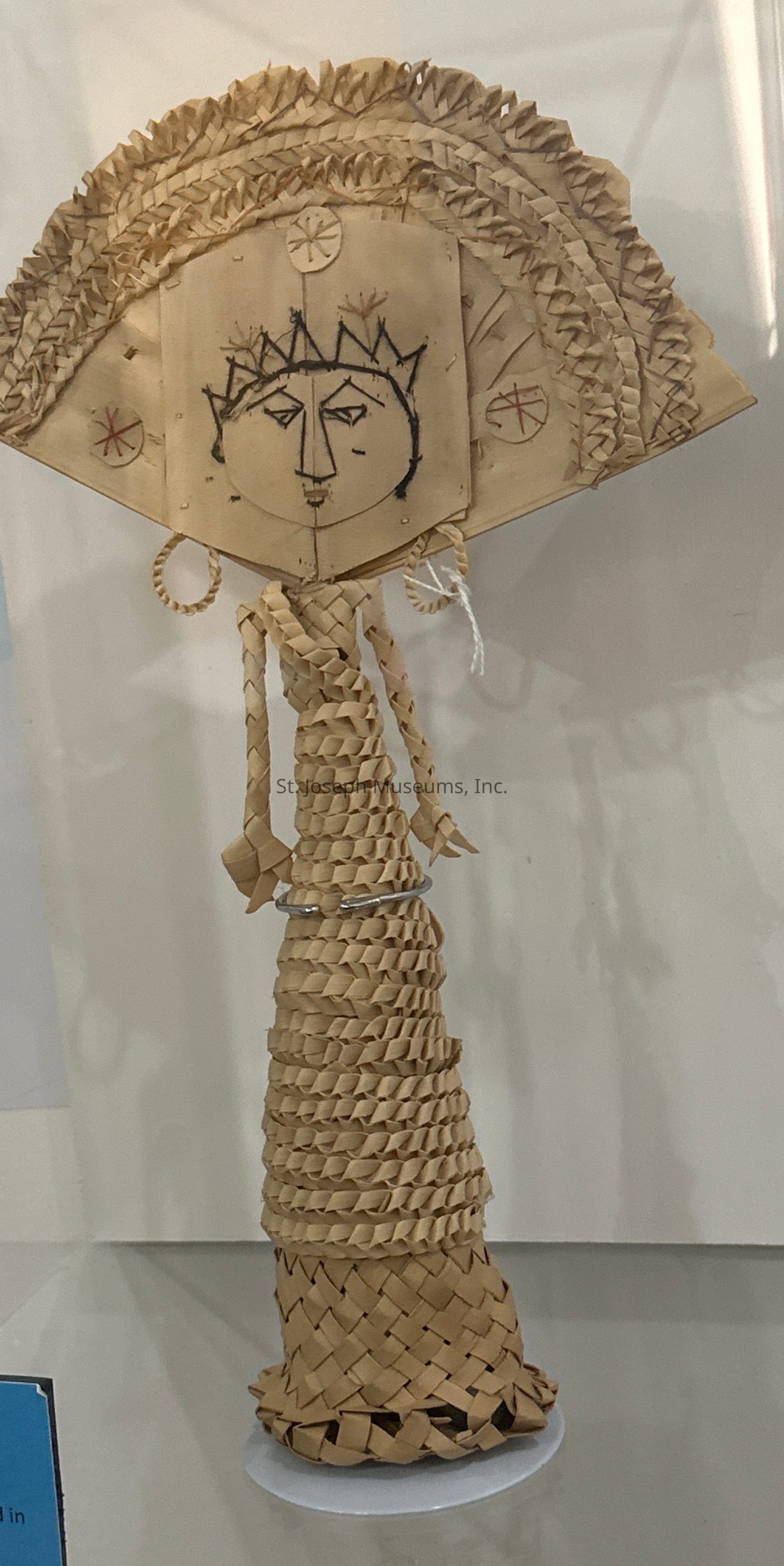 The image shows a handcrafted figure made from woven materials, possibly straw or similar fibers, featuring an artistic representation of a face. The face, drawn on a flat, square section, is adorned with geometric patterns and symbols, including a crown-like design. The figure's body is intricately braided, gradually tapering down to a rounded base. Two braided loops hang from beneath the face, likely representing arms. Above the face, layers of tightly woven material fan outwards, adding textured detail to the figure's head, resembling a headdress or hair. The entire sculpture is displayed on a flat, circular white stand, ensuring stability and focus on its artisanal construction.