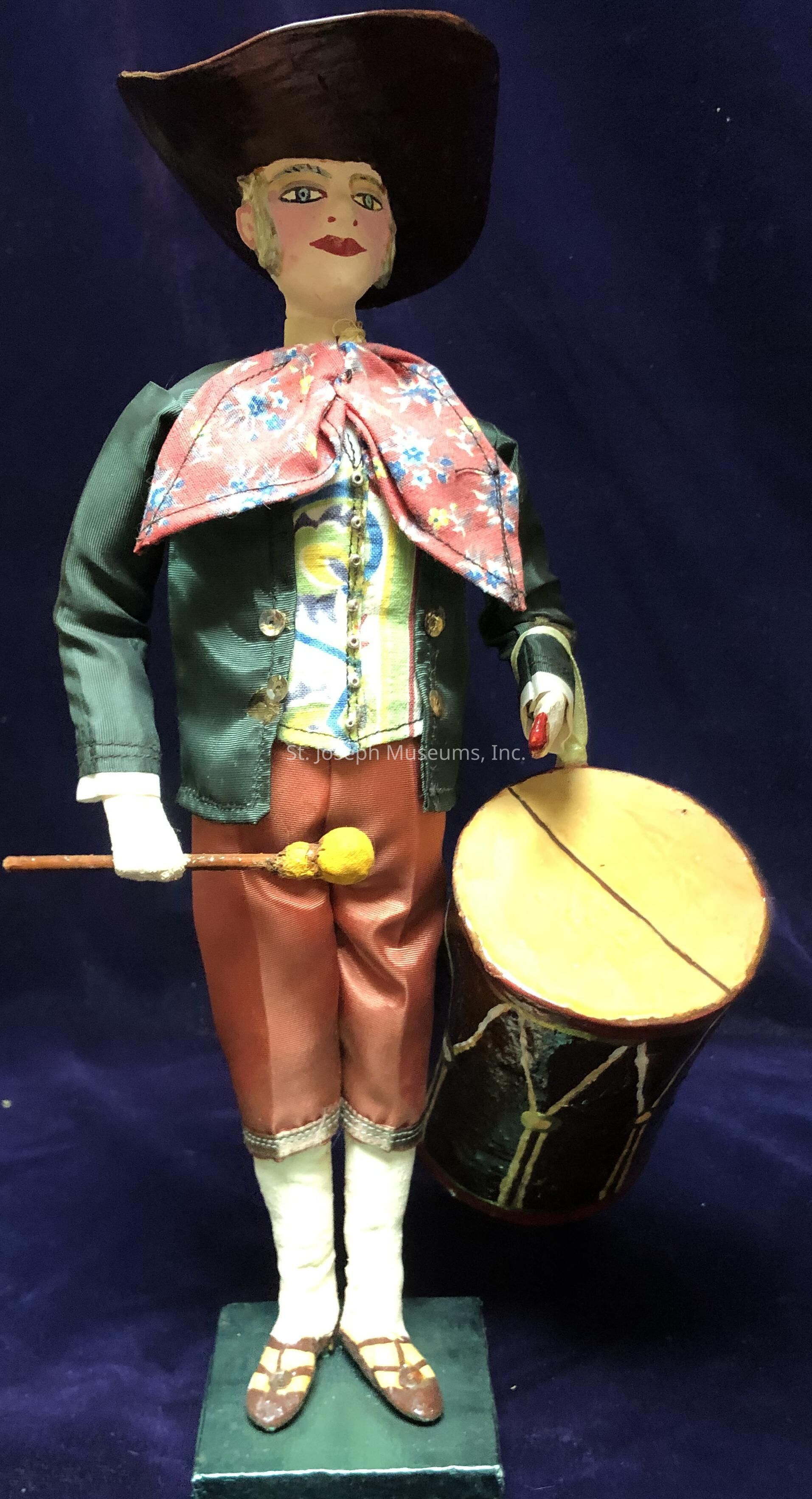 The image depicts a crafted doll designed to resemble a drummer from the historical period, standing against a rich, dark blue backdrop. The doll wears an elaborate outfit consisting of a dark green jacket with gold buttons, underneath which is a patterned vest decorated with floral and abstract designs in muted tones. Around the neck, a large, decorative red scarf with blue flowers is tied, adding a vibrant touch to the ensemble. The figure is adorned with a wide-brimmed, dark brown hat that complements the traditional attire. The attire is completed by brown pants rolled just below the knee, revealing the doll’s white socks and brown shoes, which are detailed with straps. In its right hand, the doll holds a yellow-tipped drumstick, poised over a tan-colored drum supported at its side. Each detail, from the doll's carefully painted facial features to the precise folds in its clothing, accentuates the craftsmanship dedicated to creating a historical representation through this miniature figure.