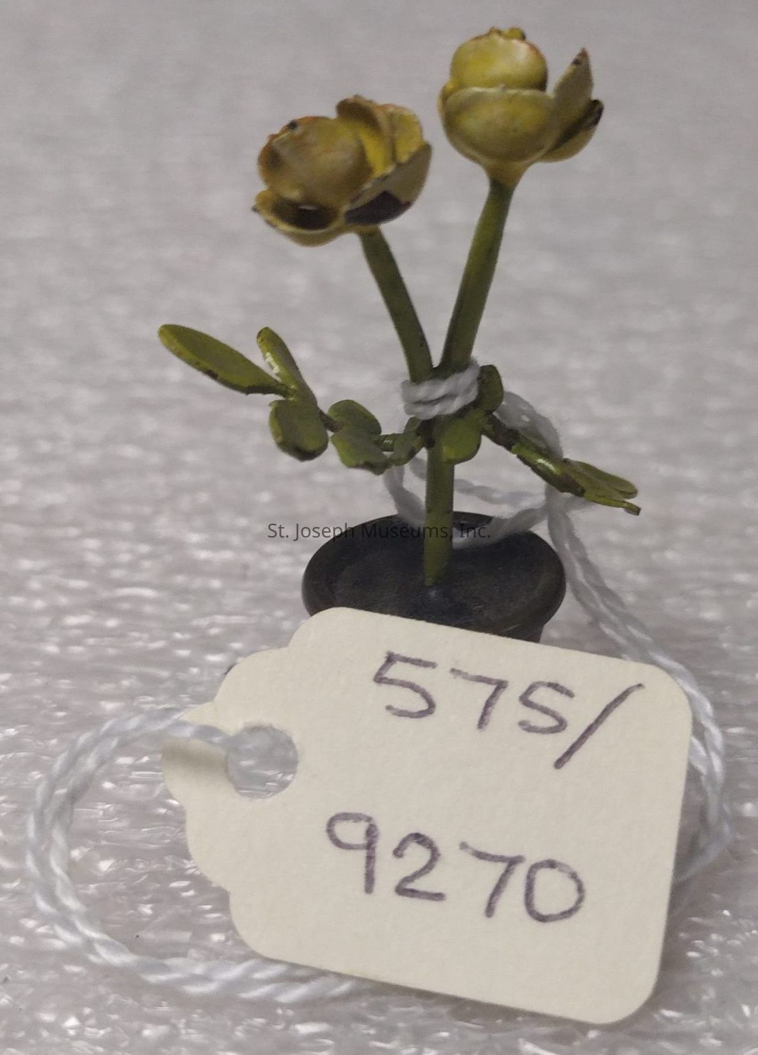 The image depicts a miniature potted plant with two stems, each topped with an intricately crafted yellow flower, emerging from an olive-green base with small leaves, all carefully tied with a piece of white thread, while attached to the pot with a small white tag featuring the numbers "575/9270" handwritten in black ink. The background is a textured white surface, which contrasts with the delicate details and colors of the plant, bringing attention to the craftsmanship and meticulous details of the miniature floral arrangement.