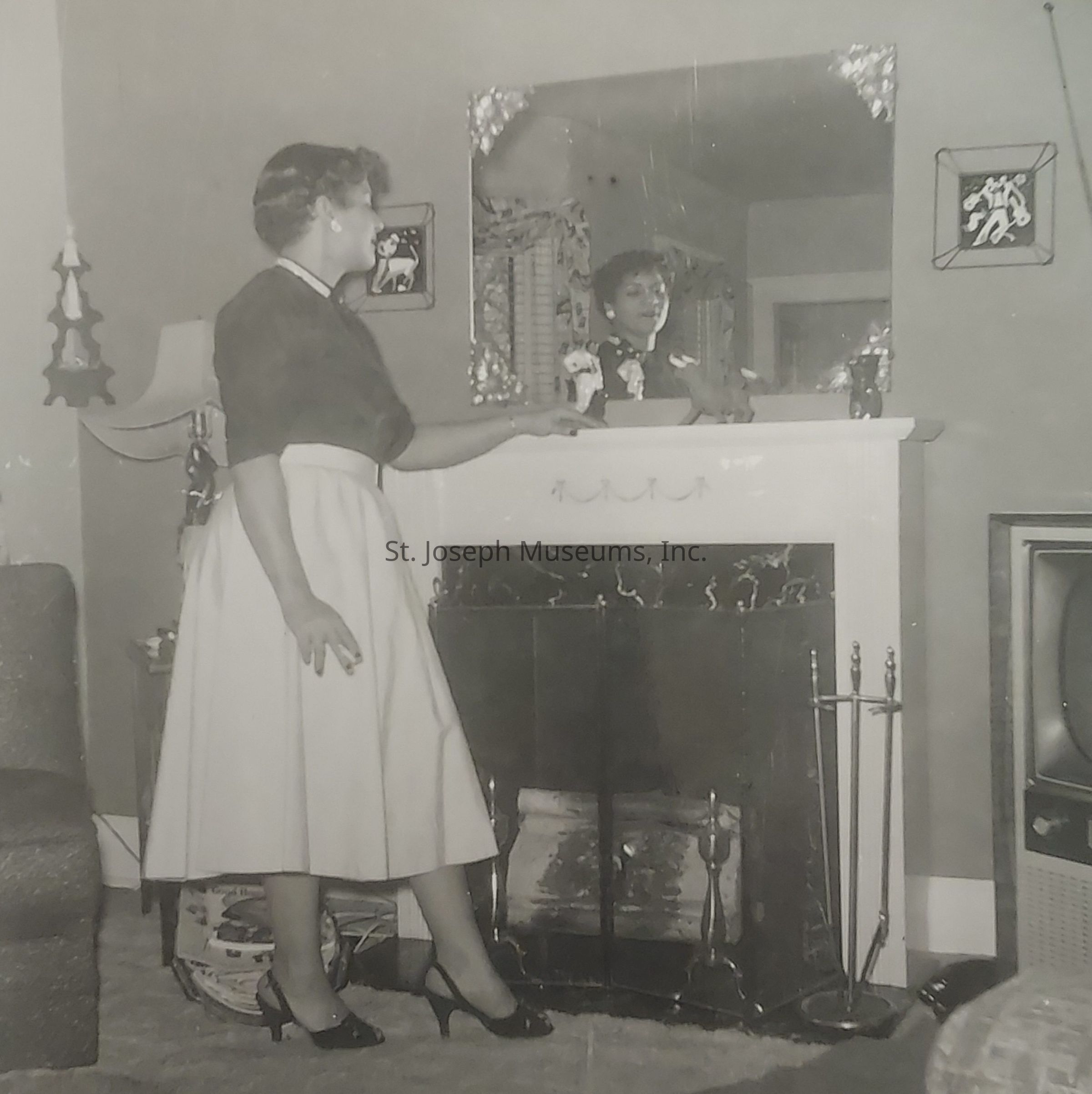 This black-and-white photograph depicts a woman standing near a fireplace in a vintage living room. She is elegantly dressed in a mid-century style outfit, consisting of a dark top and a light-colored, knee-length skirt, paired with black heels. Her hand rests on the mantelpiece, which is adorned with small decorative items. The mirror above the mantel reflects her image, further enhancing the scene’s depth. The room features period-specific decor, including a mid-century modern lamp and a television set. The setting conveys a sense of nostalgia and elegance, typical of the mid-20th century.